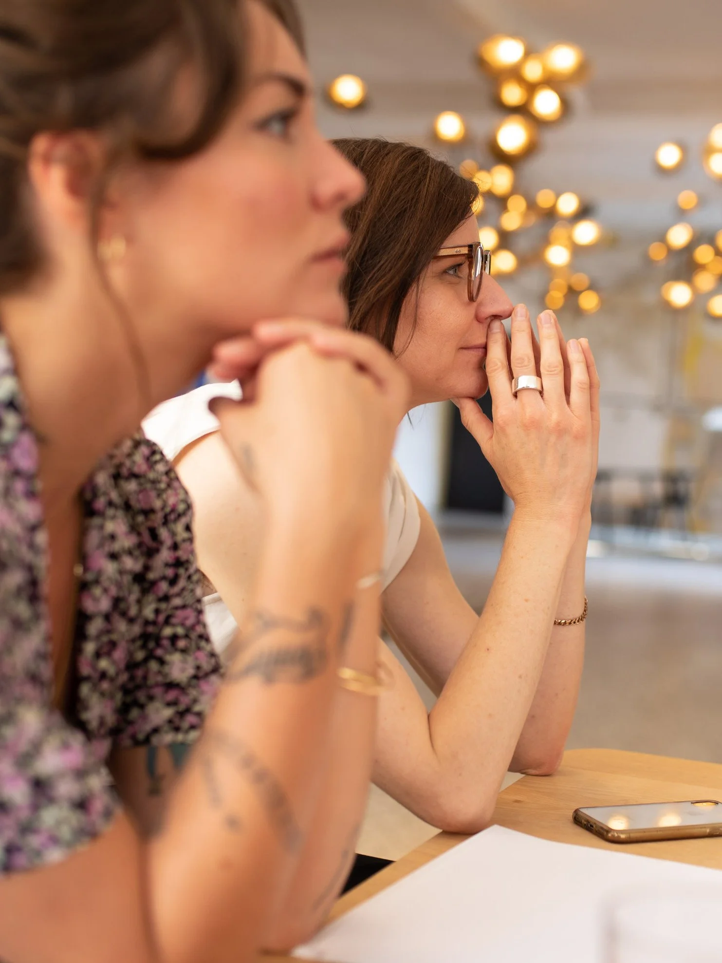 Drei Frauen sitzen nebeneinander an einem Tisch, eine schaut nach rechts, die andere trägt Brille und hat die Hand vor der Nase, im Hintergrund leuchten lichter.