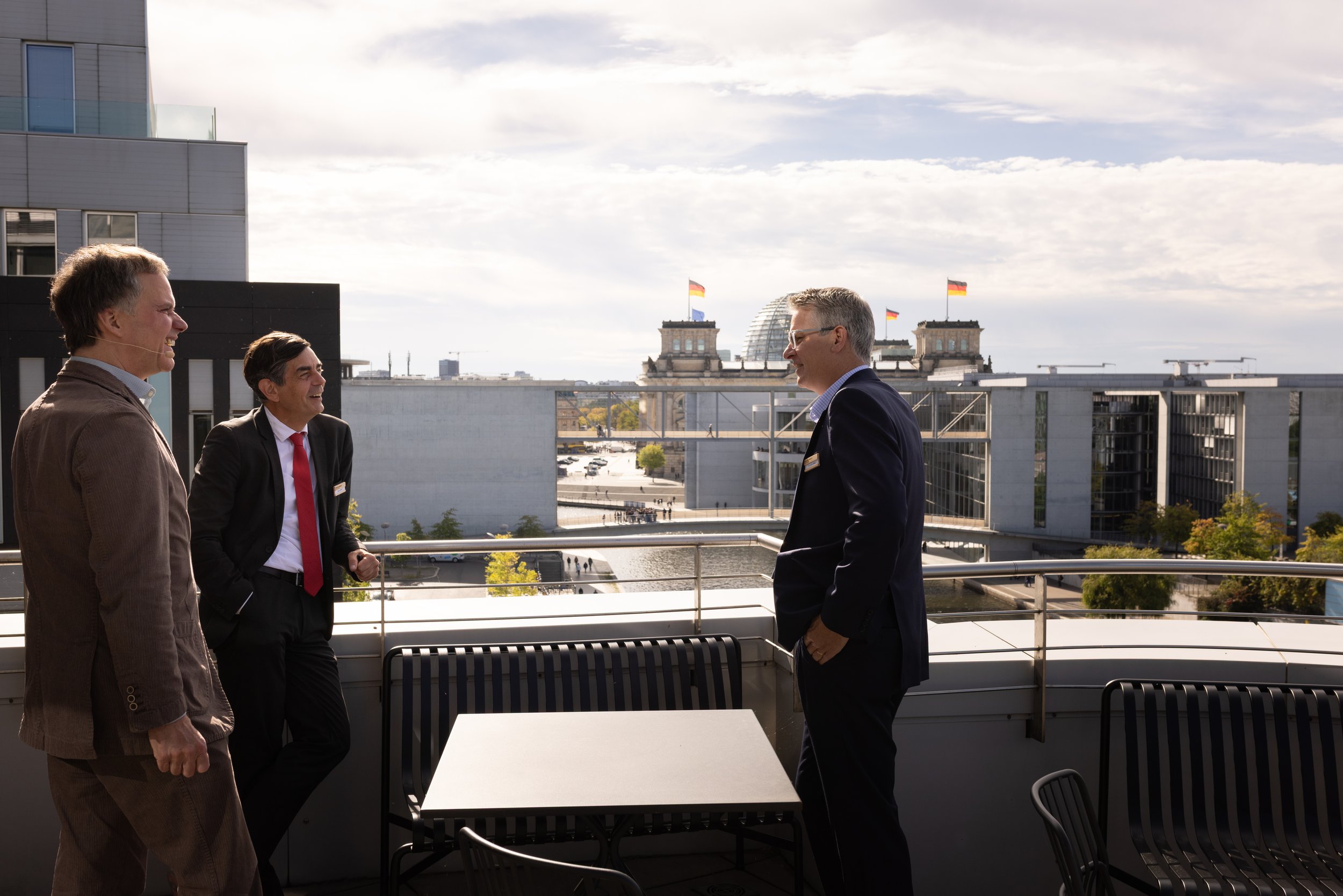 Drei Männer unterhalten sich auf einer Dachterrasse in Berlin, im Hintergrund das Deutsche Bundestag Gebäude mit deutschen Flaggen.