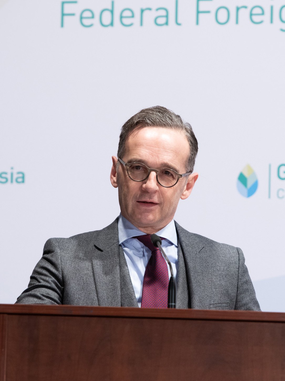 Außenminister Heiko Maas trägt einen grauen Anzug, eine blaue Hemd und eine rote Krawatte, steht hinter einer Holzpult und spricht bei einer offiziellen Veranstaltung mit einem Mikrofon. 