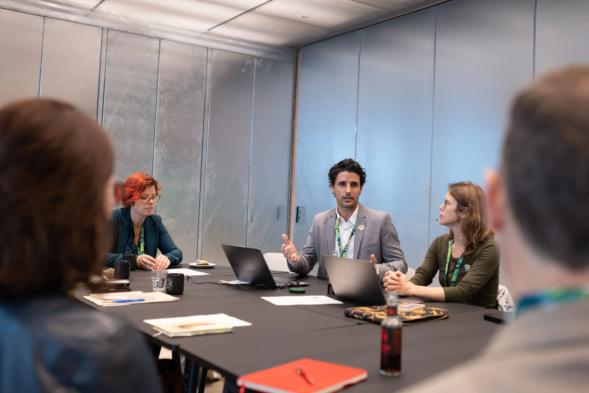 Eine Gruppe Menschen sitzt an einem Konferenztisch im Gespräch, einem Mann in einem grauen Anzug, der vor ihnen spricht.