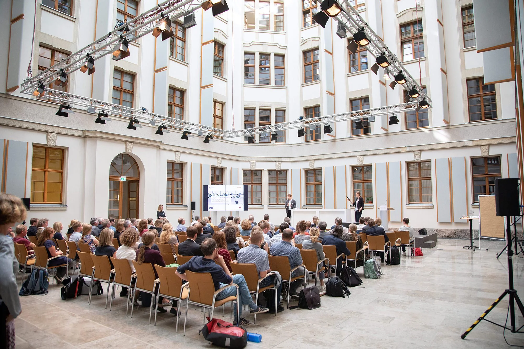 Eine große Gruppe von Menschen sitzt in einem großen Innenhof und hört einer Präsentation zu, bei der auf einem Bildschirm ein Bild gezeigt wird. Der Raum hat hohe Fenster und helle Wände.