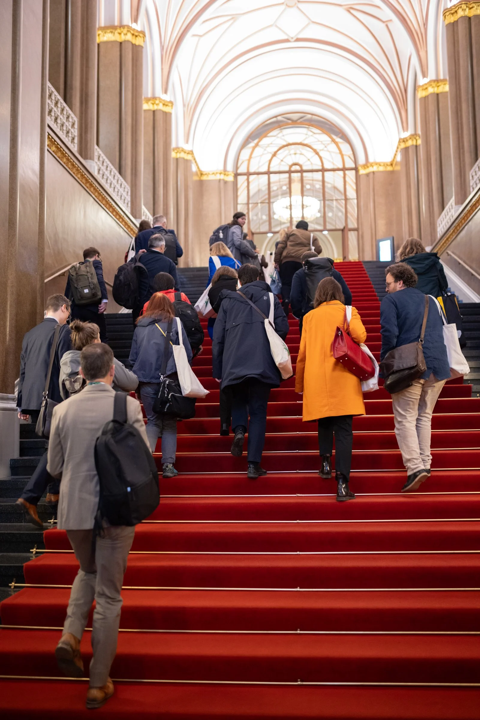 Menschen gehen die Treppe in einem prächtigen, hell erleuchteten Saal mit hohen gewölbten Decken und goldverzierten Säulen hinauf.