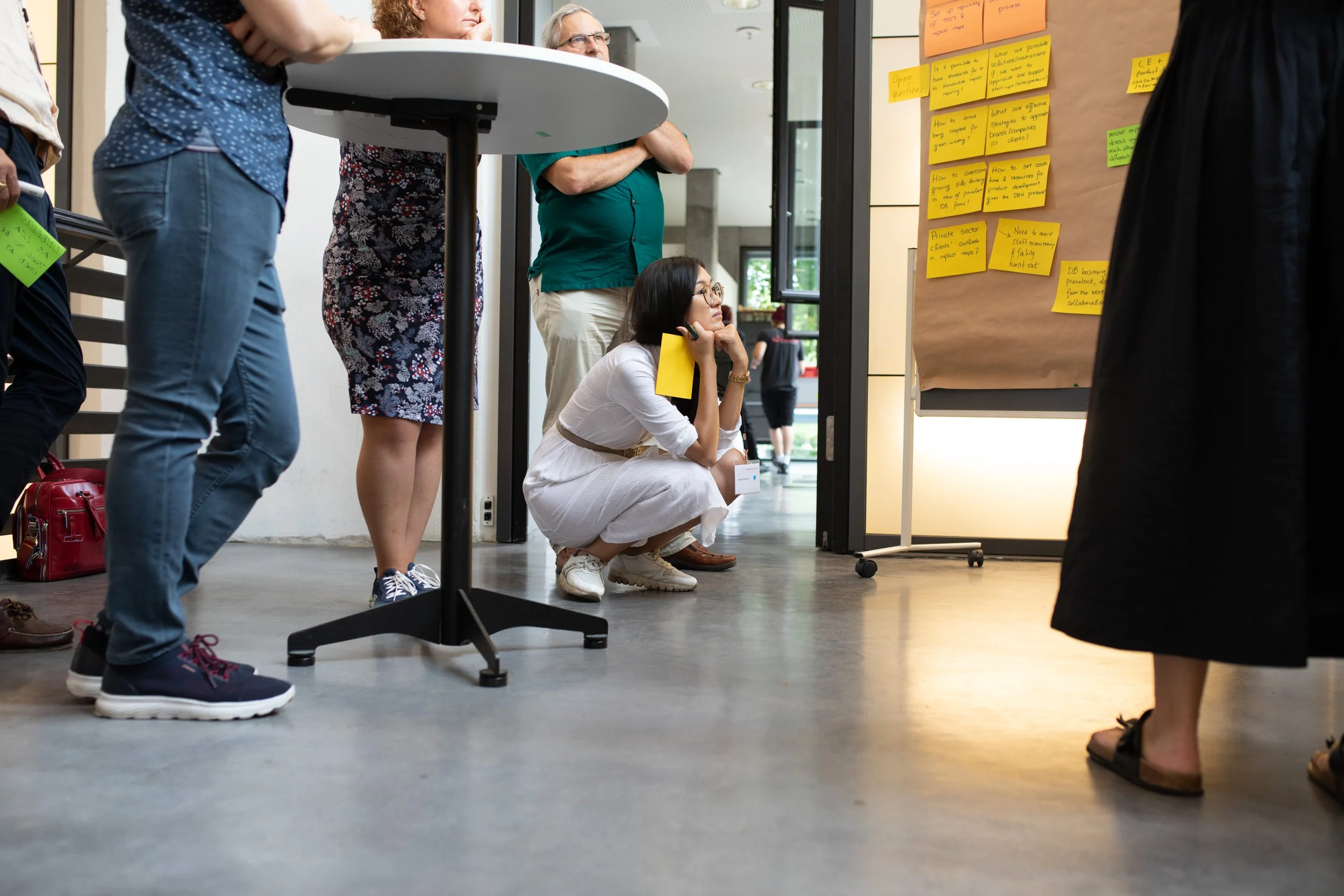 Menschen während einer Präsentation oder Diskussion in einem Raum, eine Frau sitzt auf dem Boden mit Notizblock und Stift, im Hintergrund Post-its an der Wand.