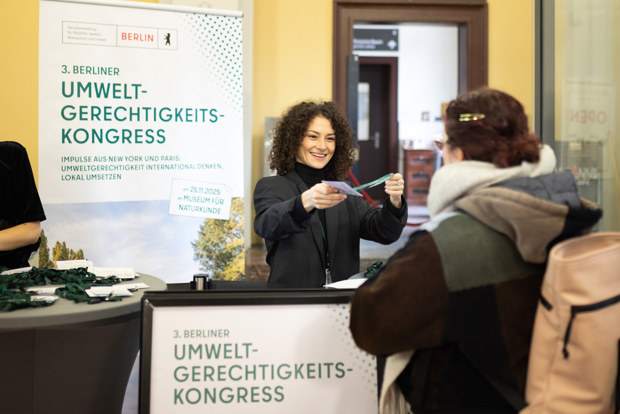 Eine Frau bei einem Umweltkongress übergibt jemandem eine Eintrittskarte. Im Hintergrund ein Poster mit dem Titel '3. Berliner Umwelt-Gerechtigkeitskongress', Datum 25.11.2025, im Naturkundemuseum.