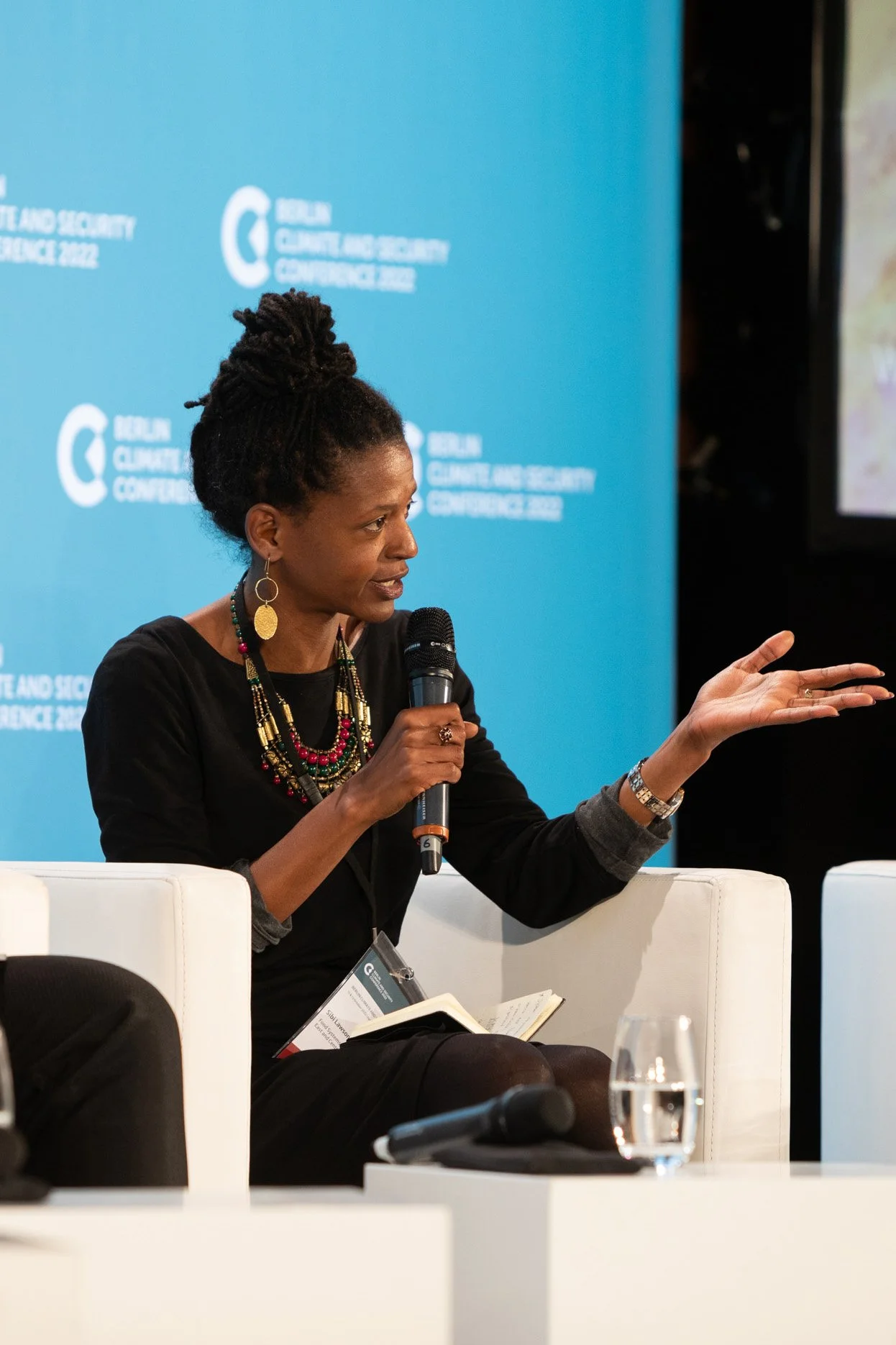 Eine schwarze Frau mit dreadlocks, die bei einer Konferenz spricht, sitzt auf einem weißen Stuhl und hält ein Mikrofon. Sie trägt bunte Ketten und große Ohrringe, im Hintergrund ein blauer Vorhang mit der Aufschrift "Berlin Climate and Security Confe