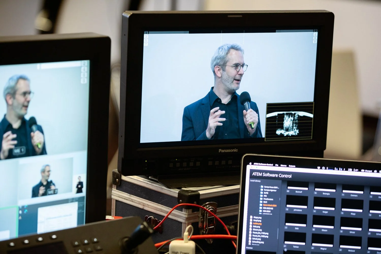 Ansicht auf eine Videoaufnahme eines Mannes, der mit einem Mikrofon spricht, auf mehreren Monitoren im Hintergrund. 