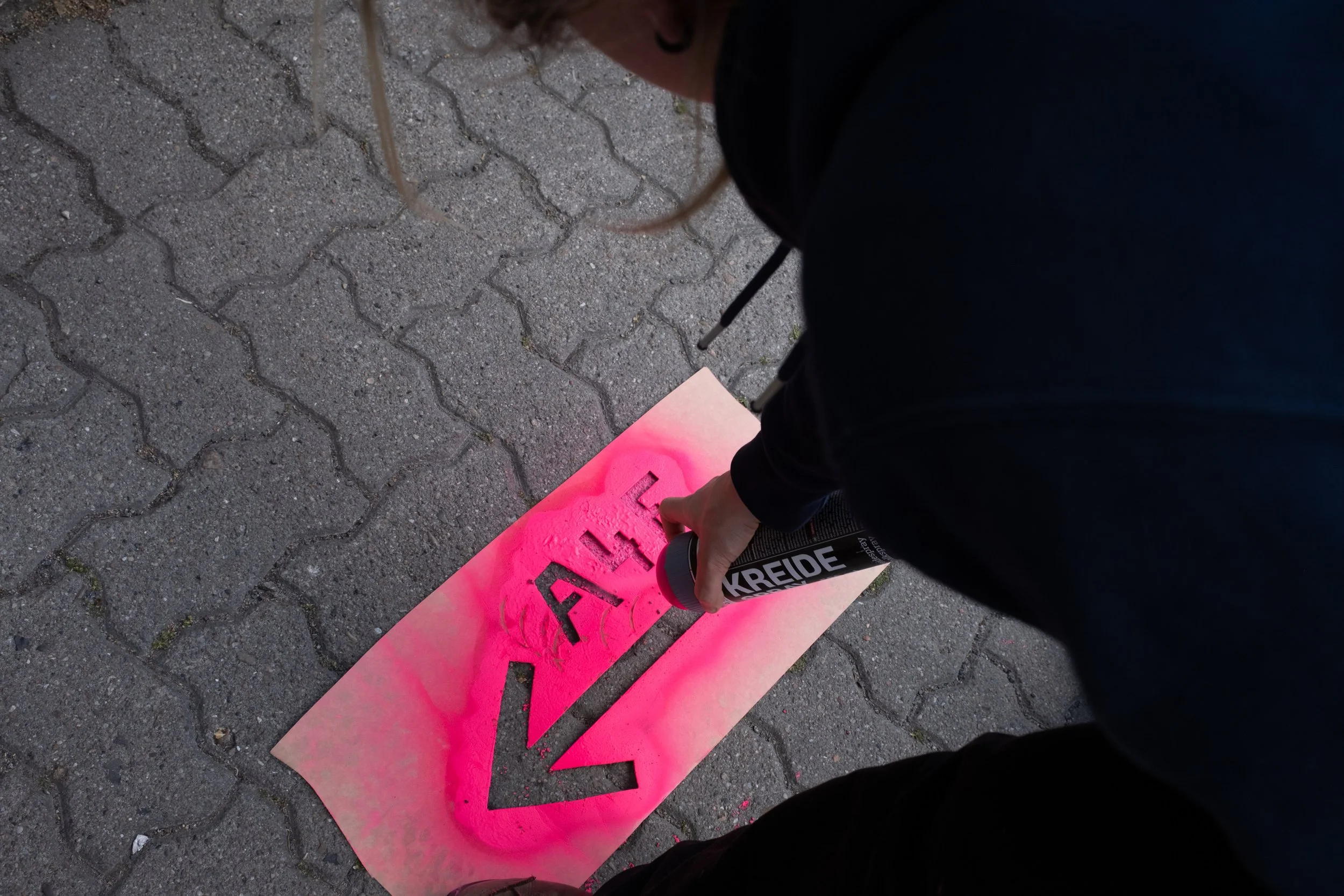 Eine Person sprüht mit einem rosa Farbspray das Wort 'GEH' auf den Boden, während sie auf einer gepflasterten Straße steht.