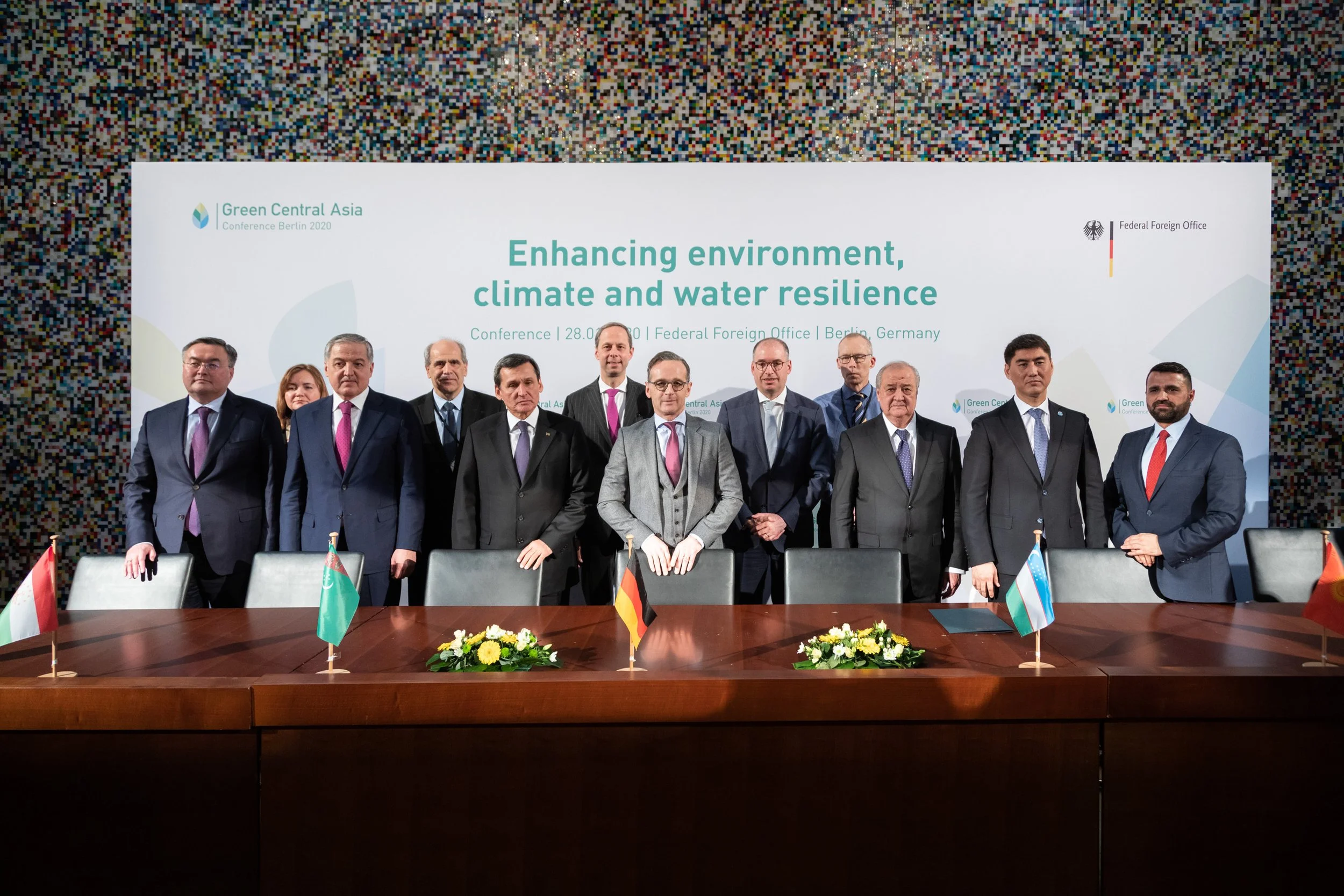 Gruppe von Männern in Anzügen bei einer Konferenz, im Hintergrund ein Bildschirm mit Text "Enhancing environment, climate and water resilience", auf dem Tisch kleine Flaggen verschiedener Länder und Blumen.