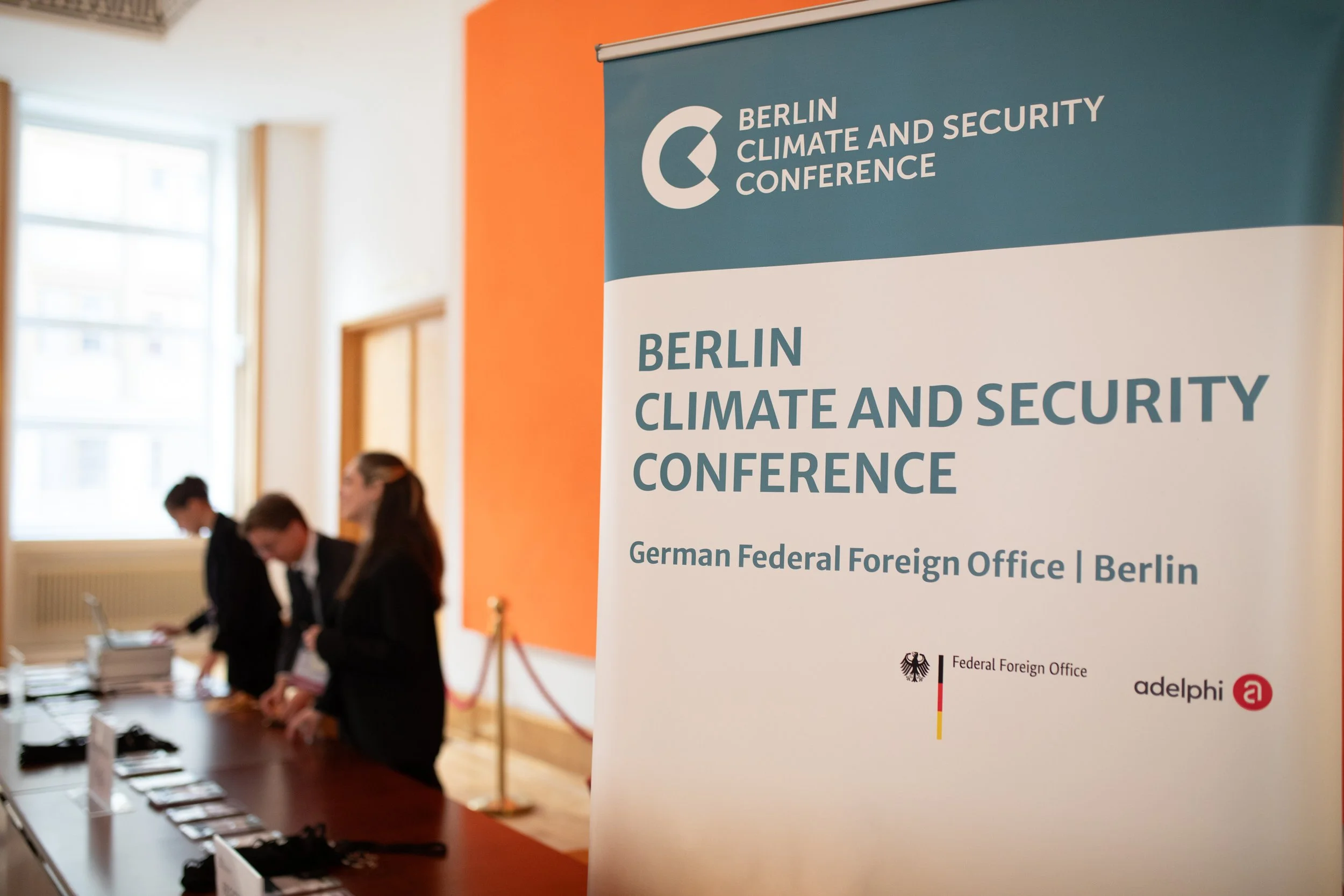 Ein Stand bei einer Konferenz mit einem großen blauen Banner, auf dem steht: "Berlin Climate and Security Conference" und "German Federal Foreign Office | Berlin". Im Hintergrund sind mehrere Personen, die an einem Tisch arbeiten.