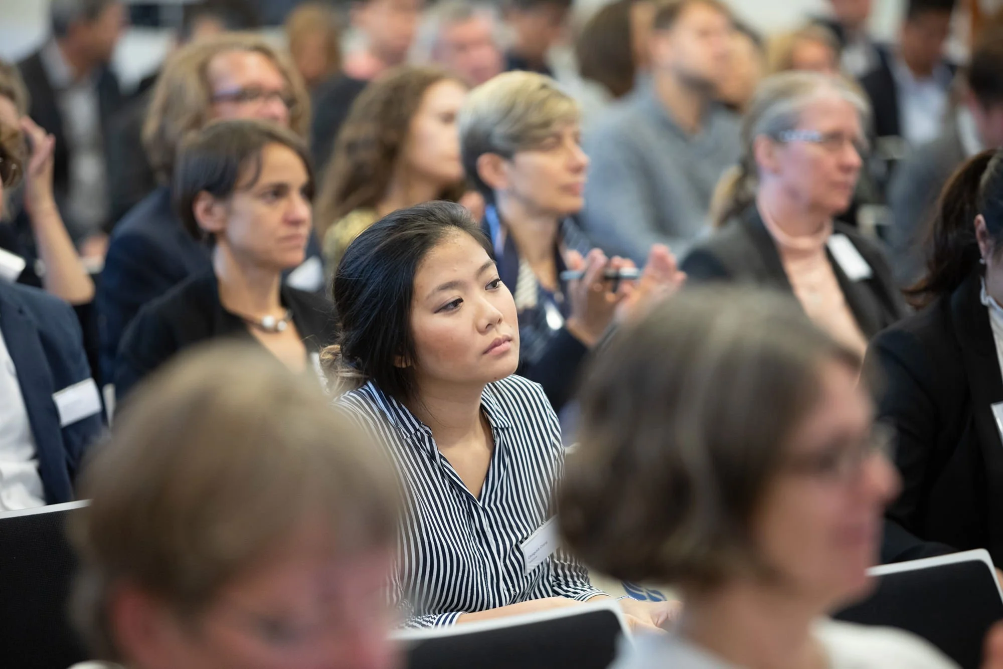 Menschen bei einer Konferenz oder einem Seminar, wobei eine junge Frau im Mittelpunkt sitzt, umgeben von anderen Zuhörern.