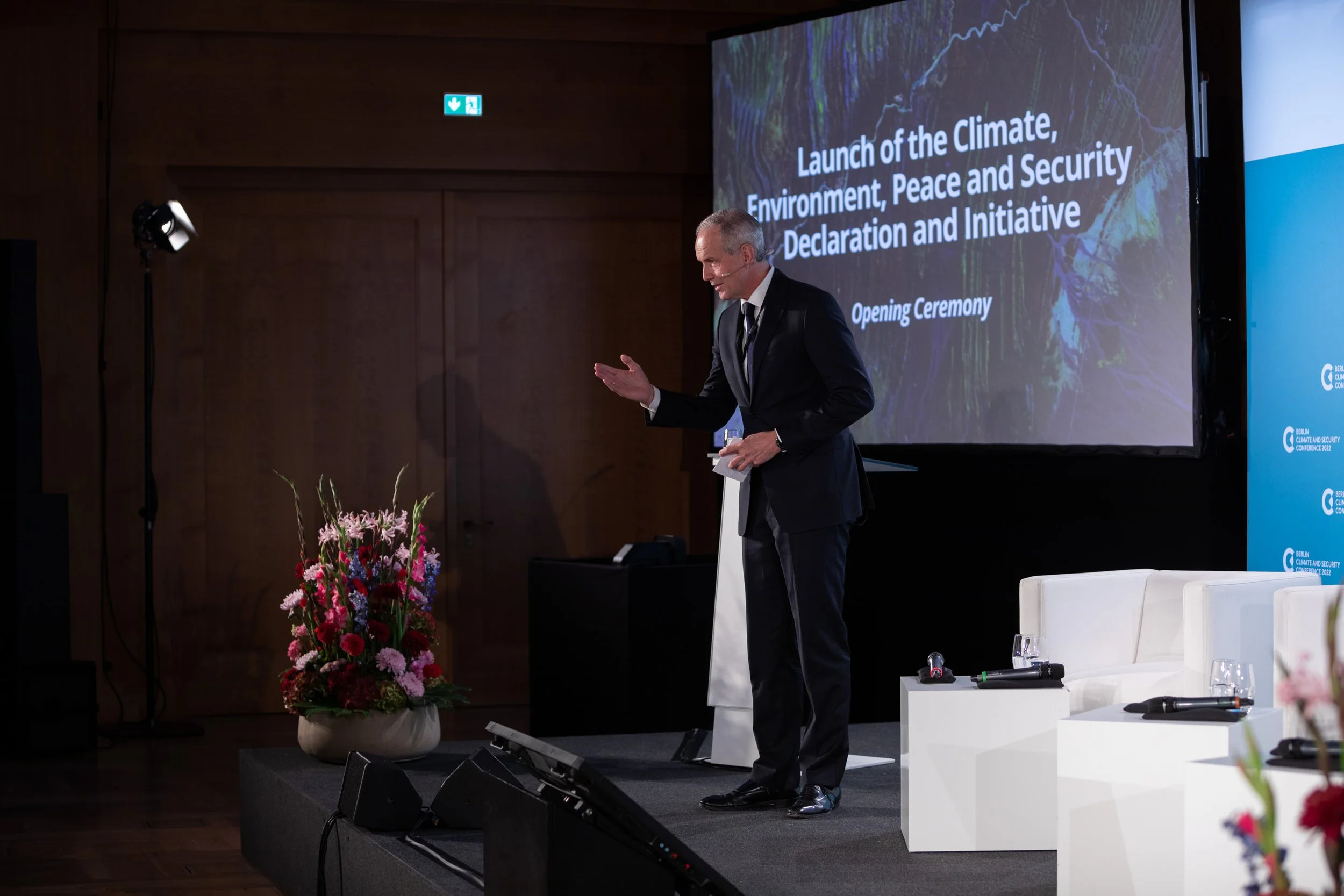 Ein Mann in Anzug spricht bei einer Konferenz vor einem großen Bildschirm mit der Aufschrift 'Launch of the climate, environment, peace and security declaration and initiative'. Im Vordergrund befinden sich Blumen, mehrere leere weiße Stühle und Mikr