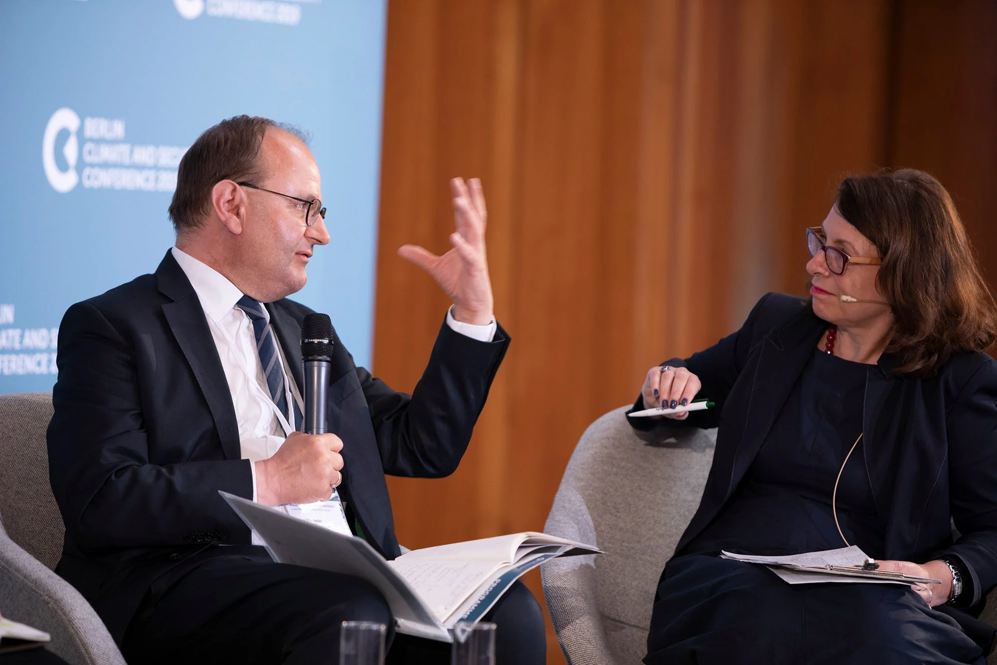 Zwei Personen bei einer Diskussion während einer Konferenz, ein Mann mit Brille, Anzug und Krawatte hält ein Mikrophon, eine Frau mit Brille und dunklem Outfit schreibt Notizen, im Hintergrund ein blauer Vorhang mit Logo.