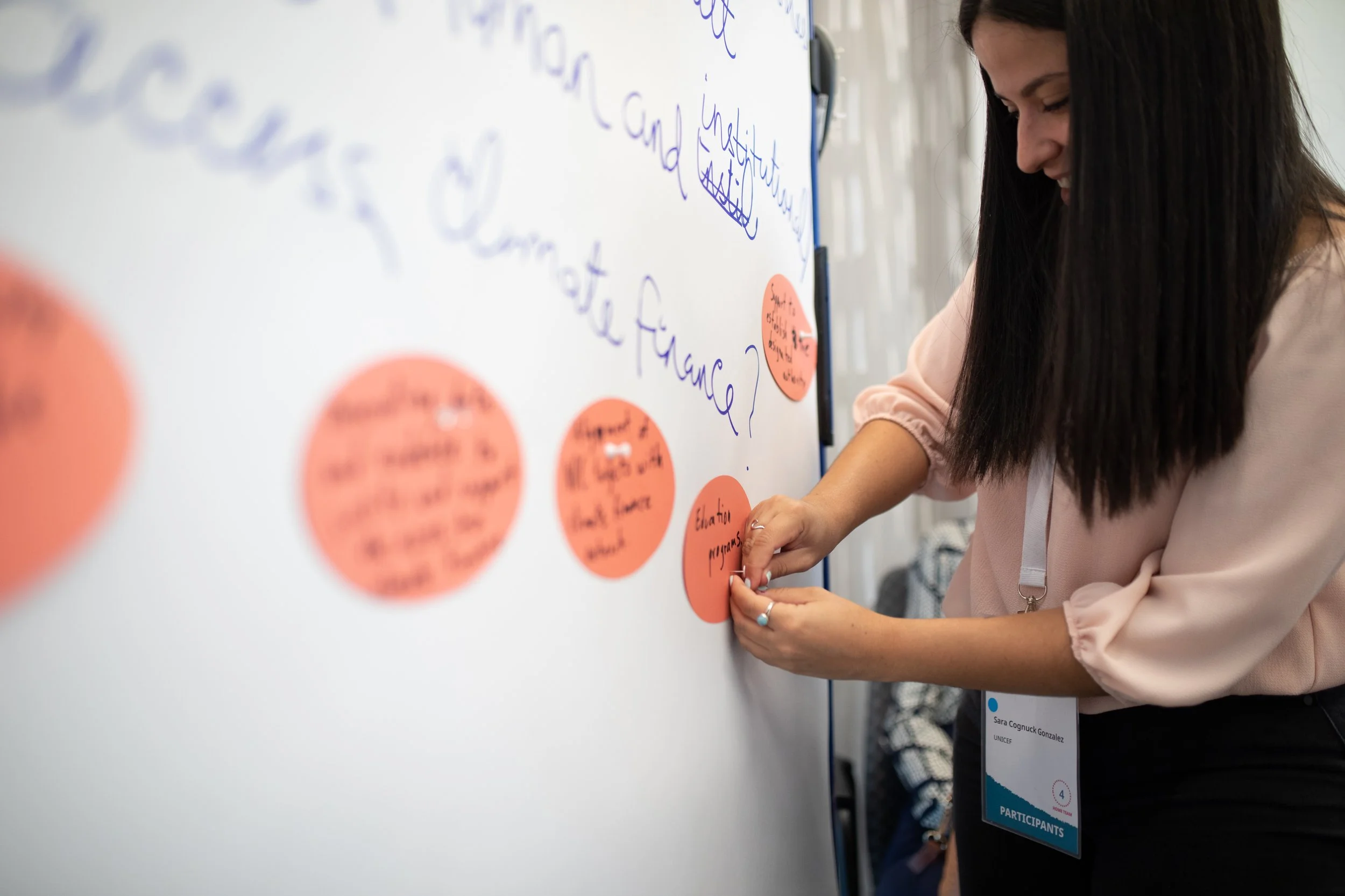 Eine junge Frau schreibt an eine Whiteboard-Tafel, die mit pinkfarbenen Post-its und blauer Handschrift bedeckt ist. Sie trägt ein Namensschild mit der Aufschrift 'Sara Cognuck Gonzalez' und ist bei einer Veranstaltung, wahrscheinlich im Bereich der 
