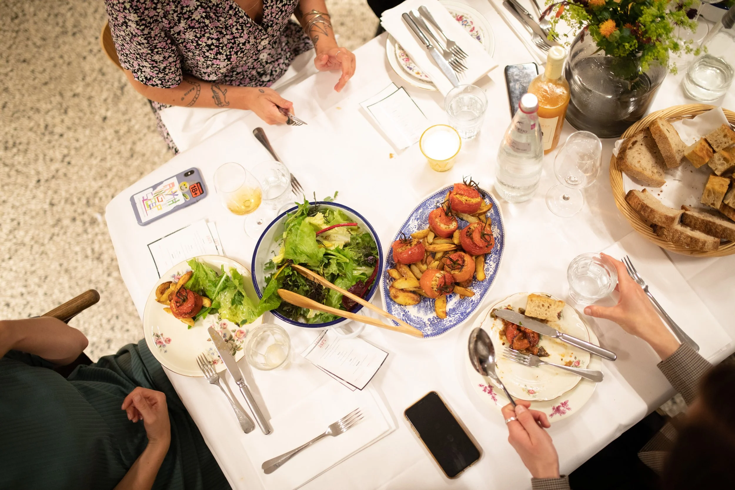 Blick auf ein gedecktes Tisch mit Salaten, Broten, Getränken und Besteck bei einem Essen im Restaurant, mit mehreren Personen, die essen und sich unterhalten.