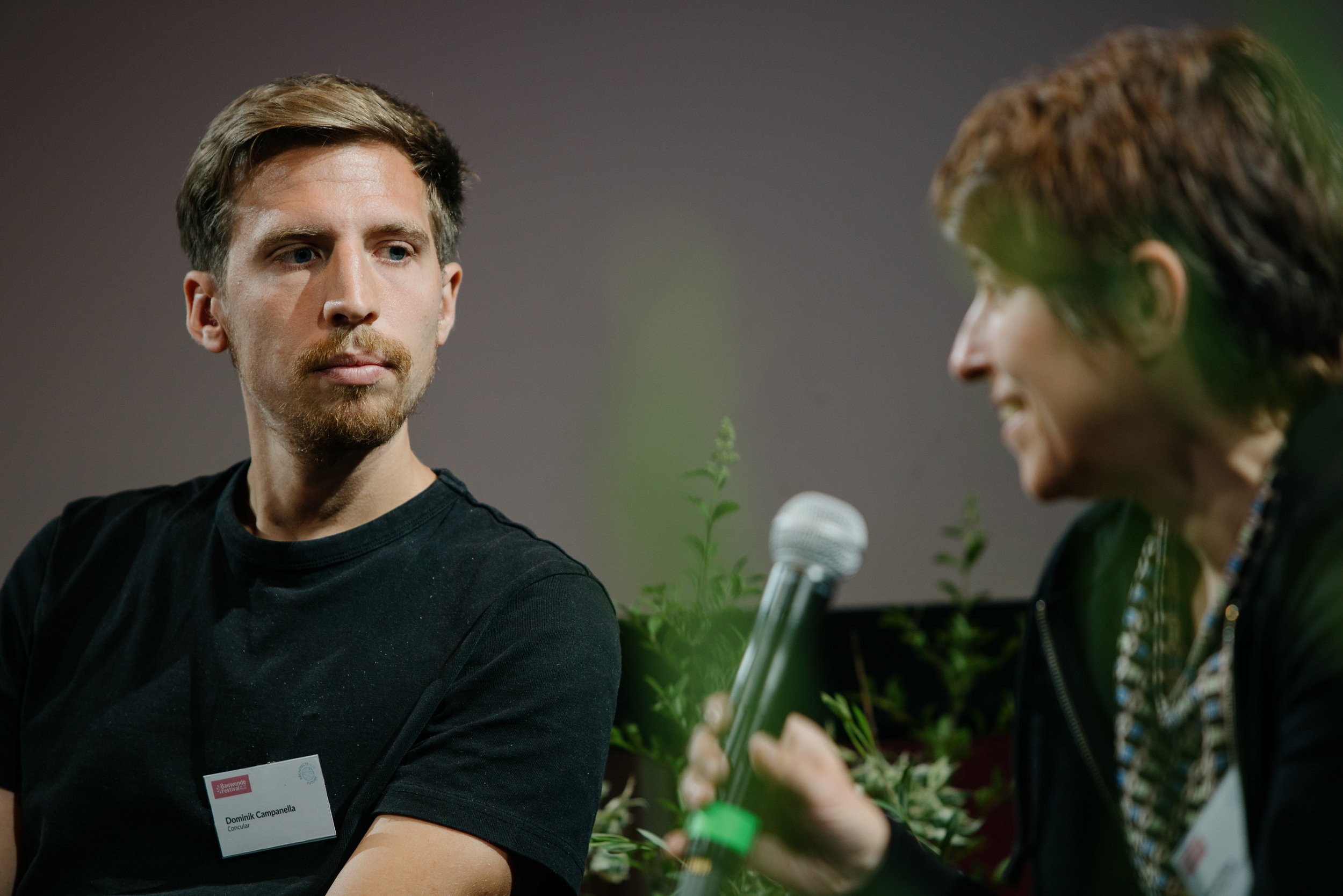 Zwei Menschen bei einer Diskussion, einer mit einem Namensschild, im Hintergrund Pflanzen, eine Person hält ein Mikrofon in der Hand.