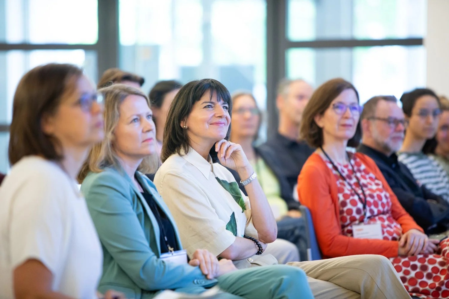 Menschen sitzen in einem Konferenzraum und hören aufmerksam einem Vortrag zu, einige notieren oder tragen Namensschilder.