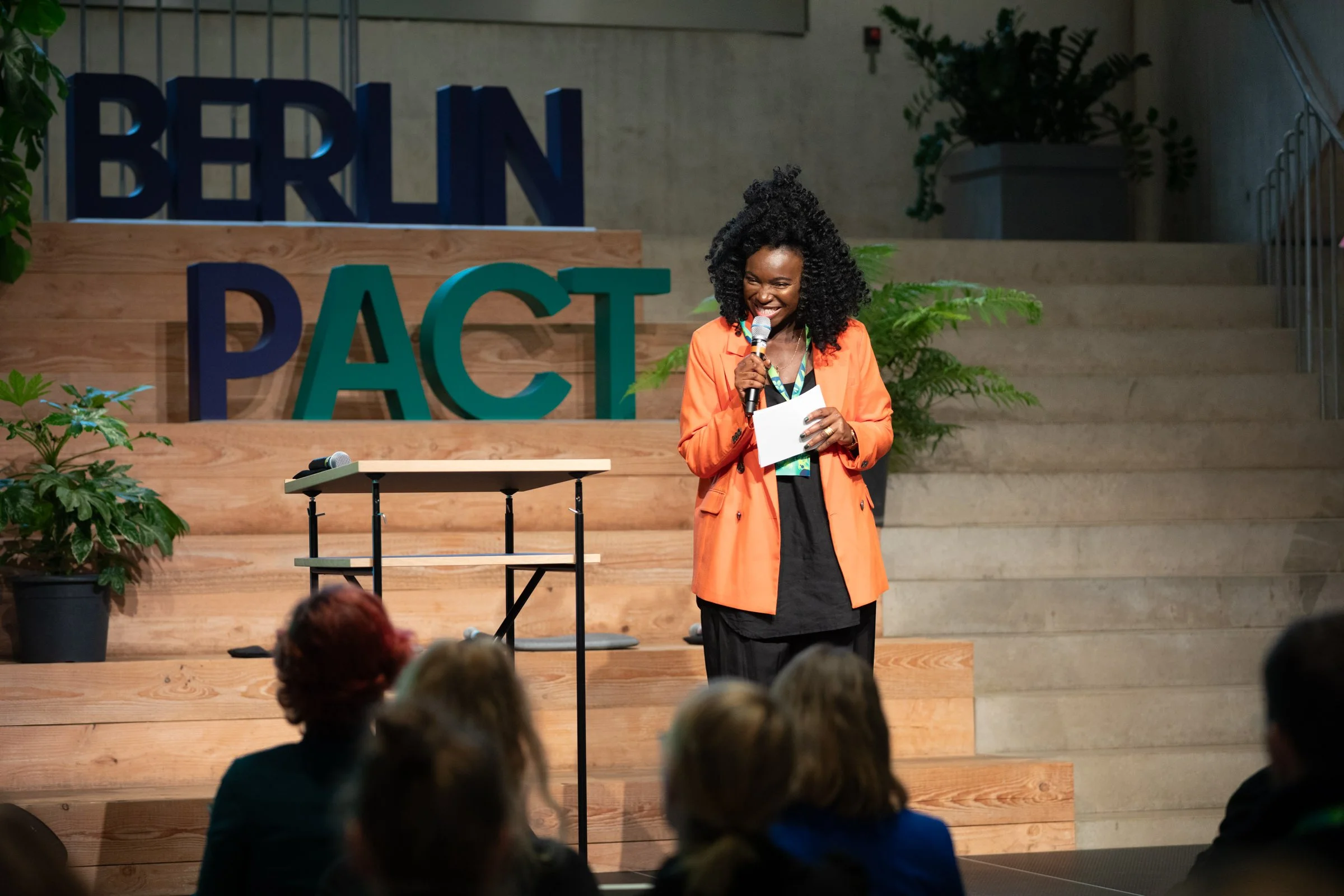 Eine Frau mit Locken, die ein Mikrofon hält und vor einem Publikum spricht, im Hintergrund steht ein großes Wort 'BERN' und 'PACT' auf einer Holzplattform, umgeben von Pflanzen