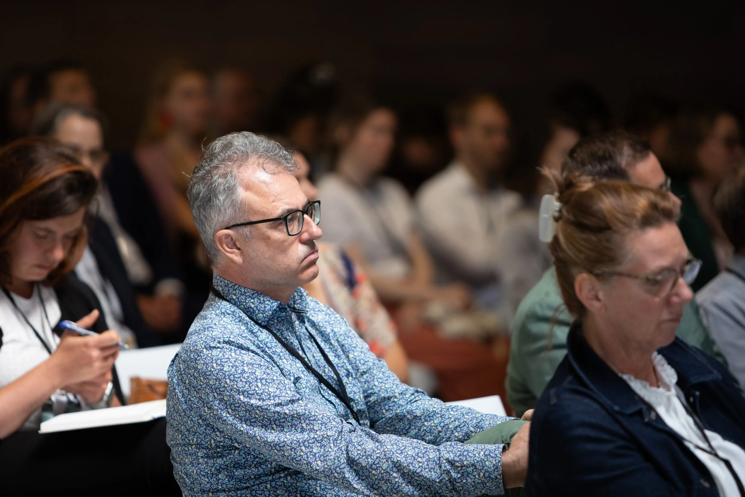 Menschen hören aufmerksam bei einer Konferenz oder Vorlesung, einige schreiben Notizen, alle sitzen in einem dunklen Raum, im Vordergrund ein älterer Mann mit Brille und einem blaugeblümten Hemd.