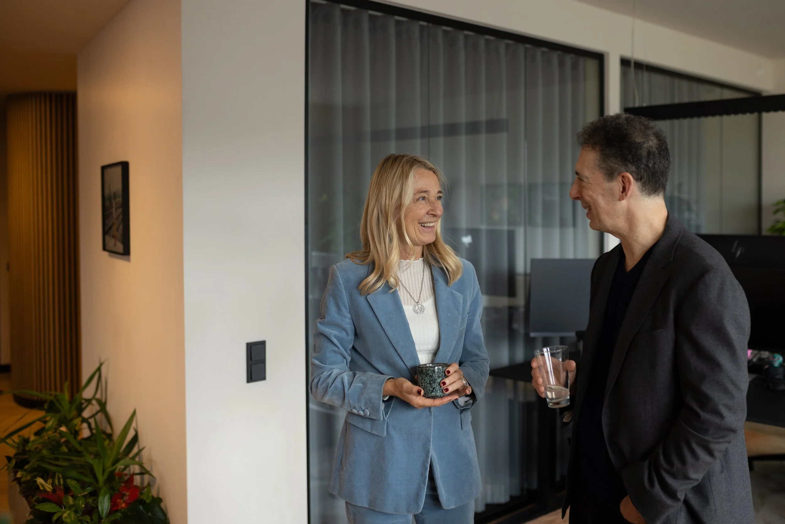 Zwei Menschen, eine Frau und ein Mann, unterhalten sich in einem modernen Innenraum. Die Frau trägt einen blauen Anzug und hält eine Tasse, während der Mann ein Glas Wasser hält. Sie lächeln beide freundlich.