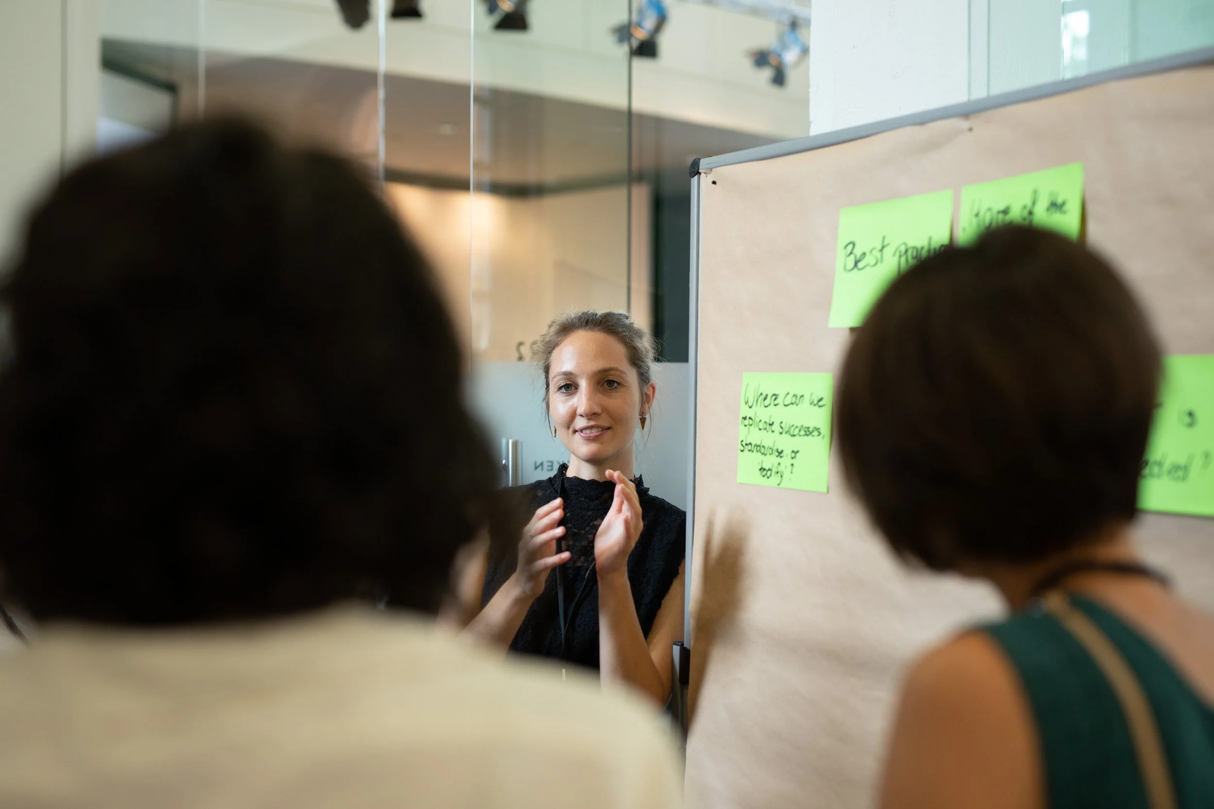 Eine junge Frau steht vor einem Whiteboard mit Post-it-Notizen und spricht zu einer Gruppe, die im Vordergrund sichtbar ist, in einem modernen Raum mit Glas und Spiegel.