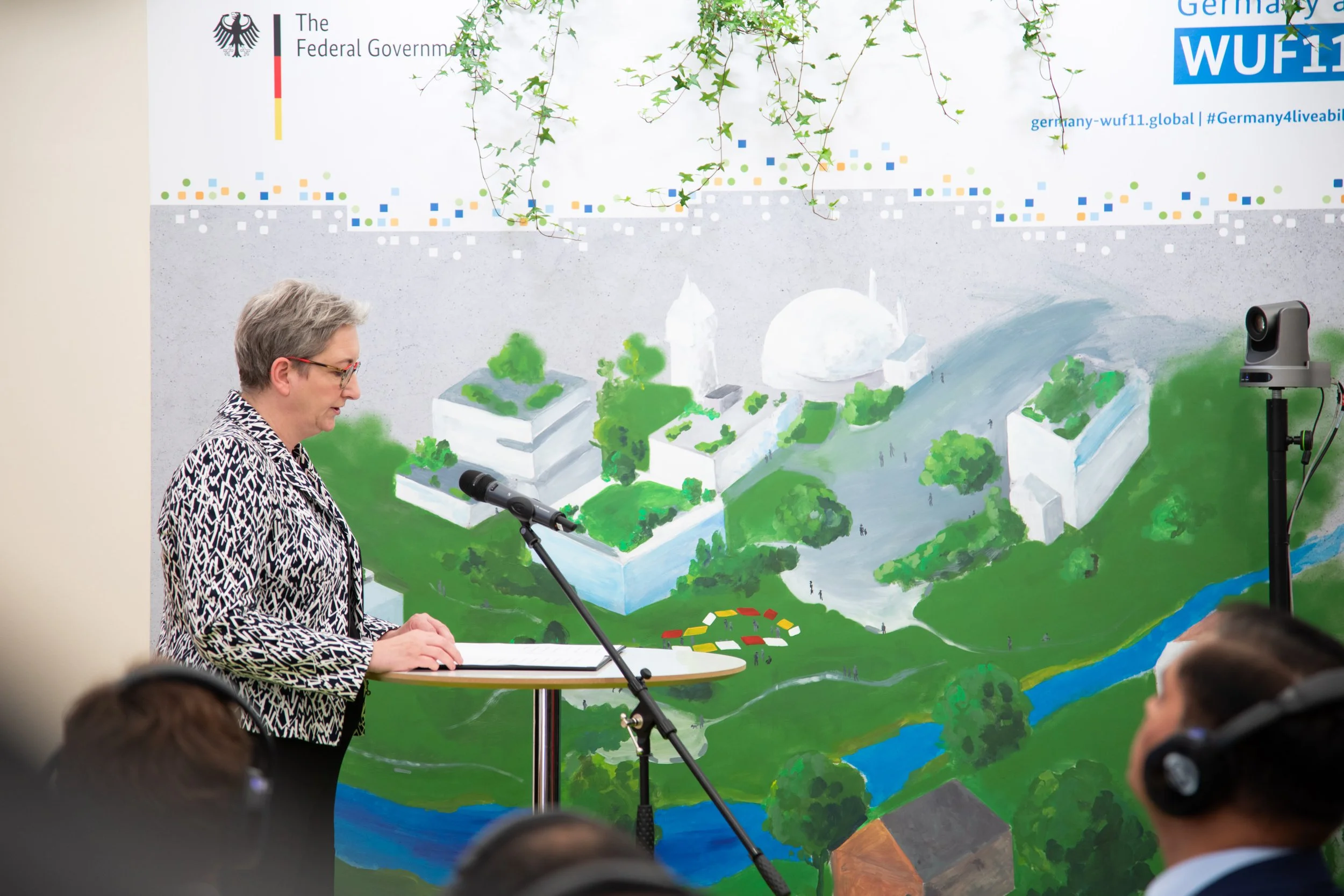 Eine Frau spricht bei einer Veranstaltung vor Publikum, im Hintergrund eine Wand mit einer Illustration eines Stadtplans und dem Text 'The Federal Government' sowie Logos und Hashtags. Die Szene ist in einem Raum mit einer Kamera auf einem Stativ zu 