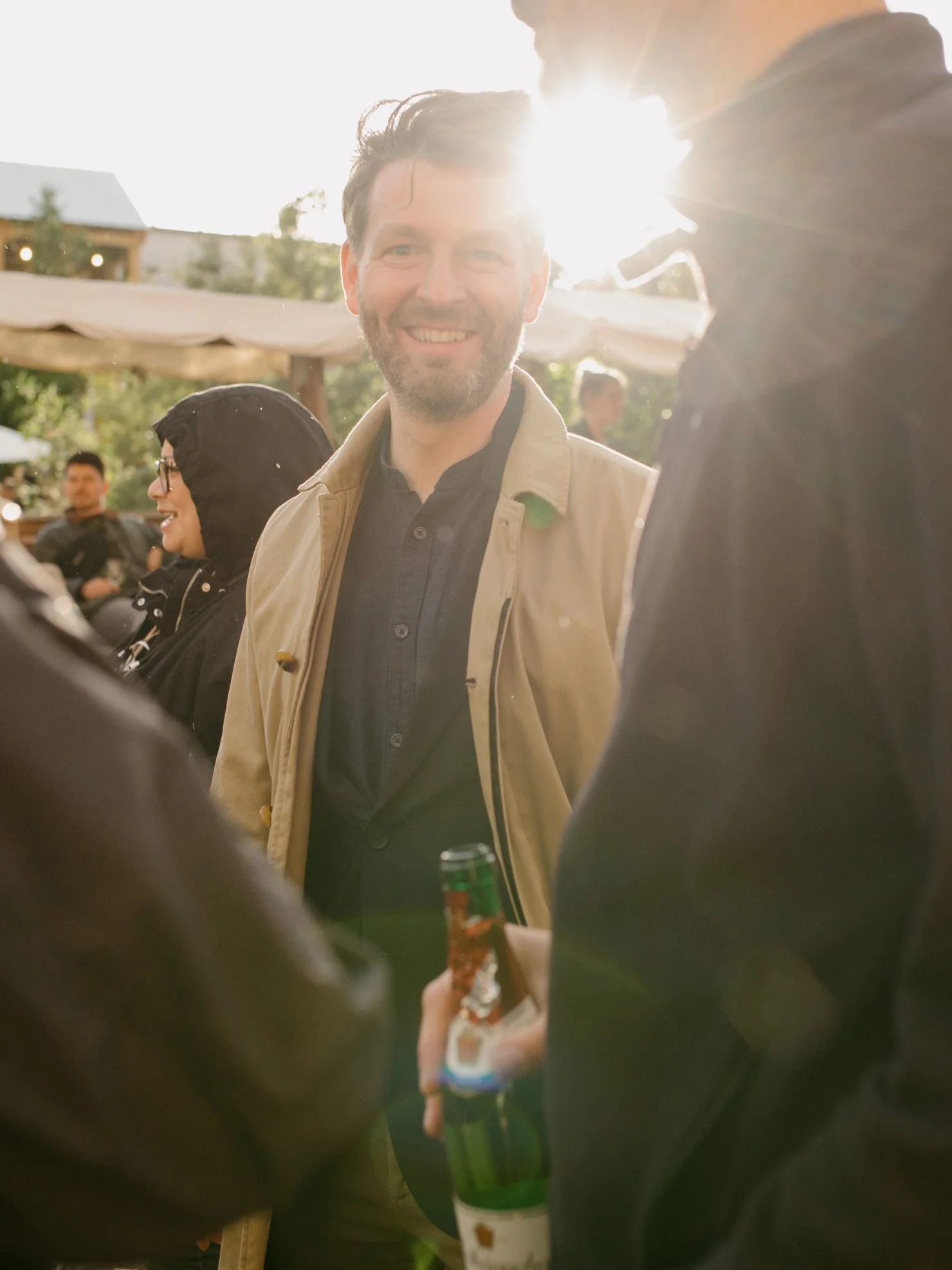 Menschen bei einem Treffen im Freien, Sonnenlicht im Hintergrund, lachender Mann im Mittelpunkt, Bierflasche in der Hand, fröhliche Atmosphäre.