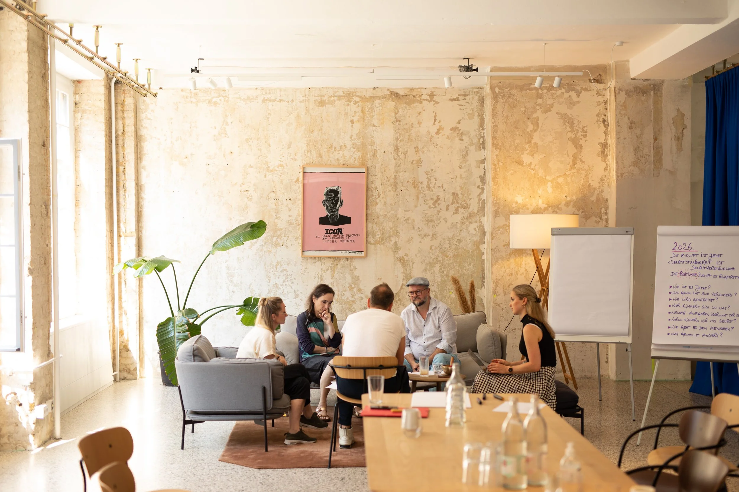Gruppe von Menschen in einer gemütlichen Lounge, die sich unterhalten, im Raum mit abblätternder Wand und Pflanzen, einen Whiteboard mit Notizen.