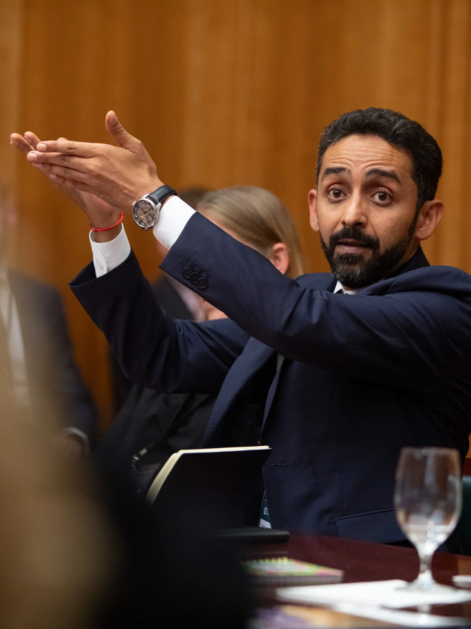 Ein Mann mit dunklem Haar und Bart, gekleidet in einem Anzug, zeigt eine Geste mit hochgehobener Hand während einer Sitzung oder Diskussion, im Hintergrund eine Person mit blonden Haaren.