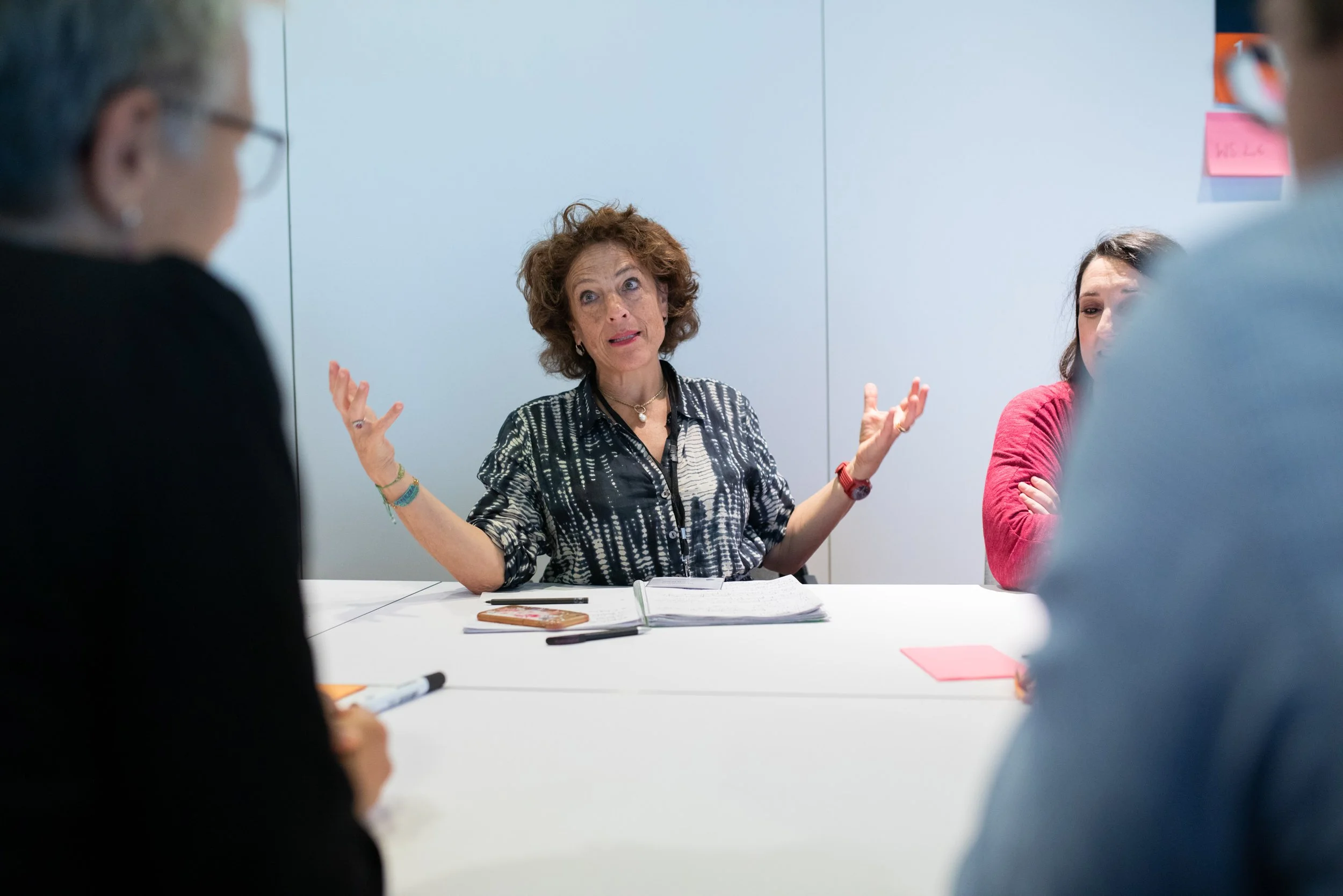Eine ältere Frau spricht während eines Meetings mit drei jüngeren Menschen in einem modernen Büro. Sie sitzt an einem weißen Tisch, hat kurze, lockige Haare, trägt eine schwarz-weiße Bluse und wirkt engagiert.