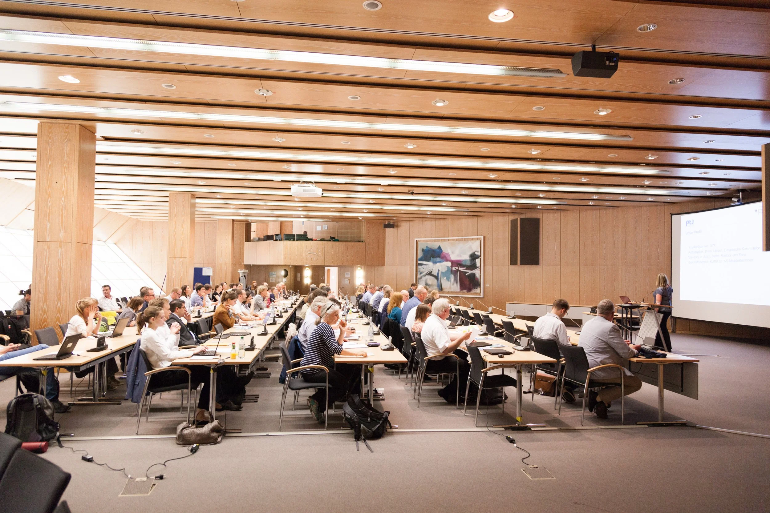 Ein großes Konferenzsaal voller Menschen, die an Tischen sitzen und einem Vortrag zuhören. Im Hintergrund ist eine große Leinwand mit Präsentationsfolien.