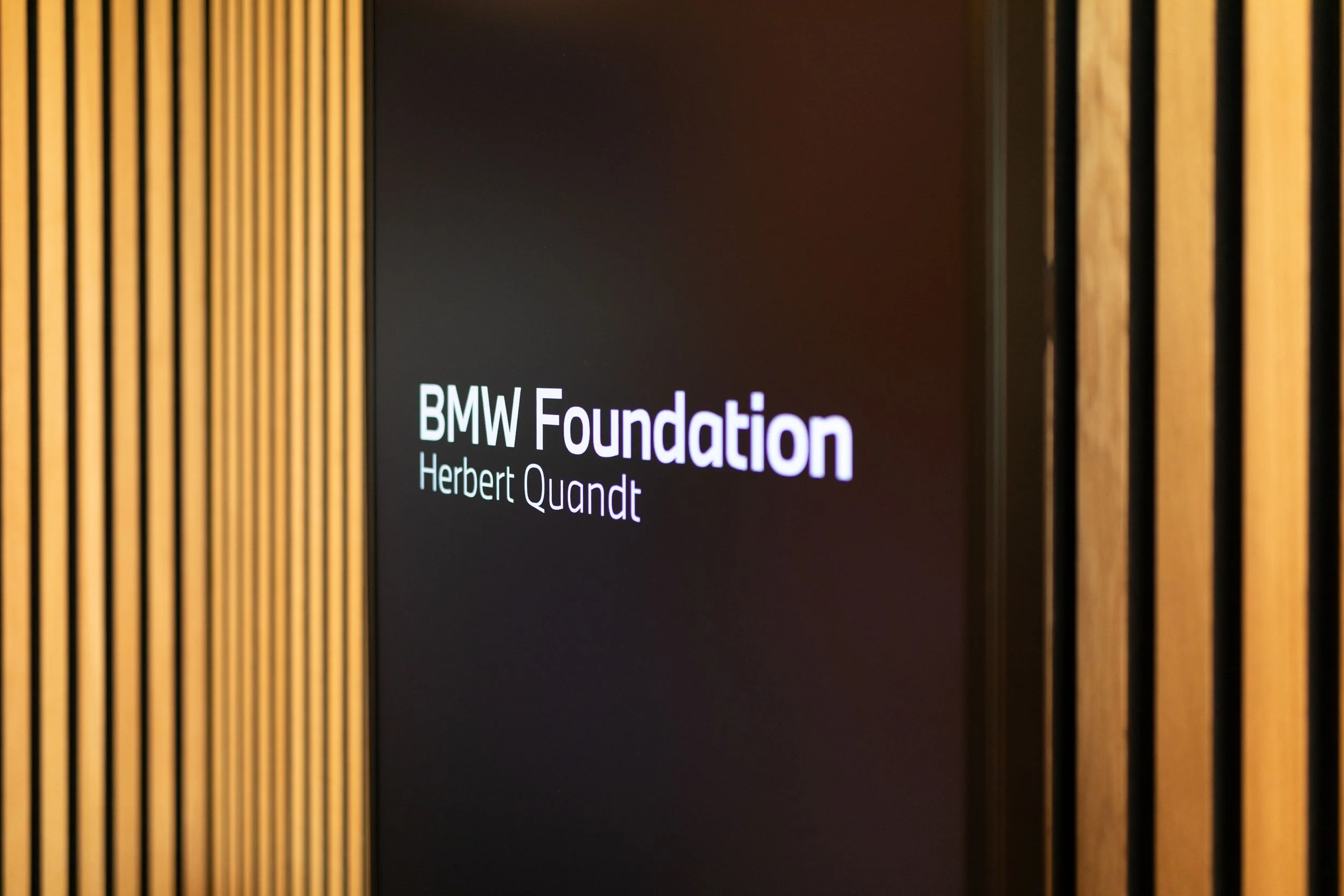 Ein Bildschirm mit dem Text 'BMW Foundation Herbert Quandt' neben vertikal angeordneten Holzlamellen.