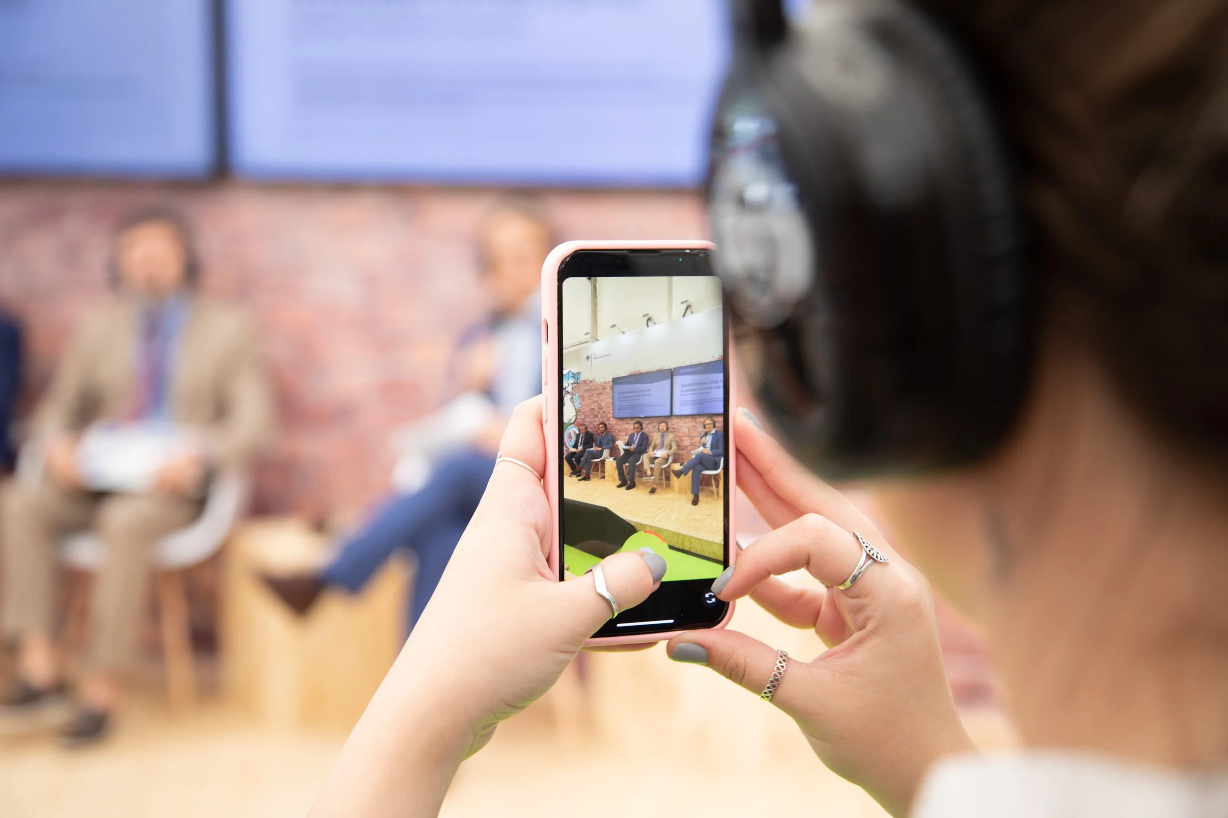 Eine Person hält ein Smartphone in der Hand und fotografiert eine Bühnenszene mit mehreren Männern, die auf Stühlen sitzen, während im Hintergrund eine Projektionswand zu sehen ist.