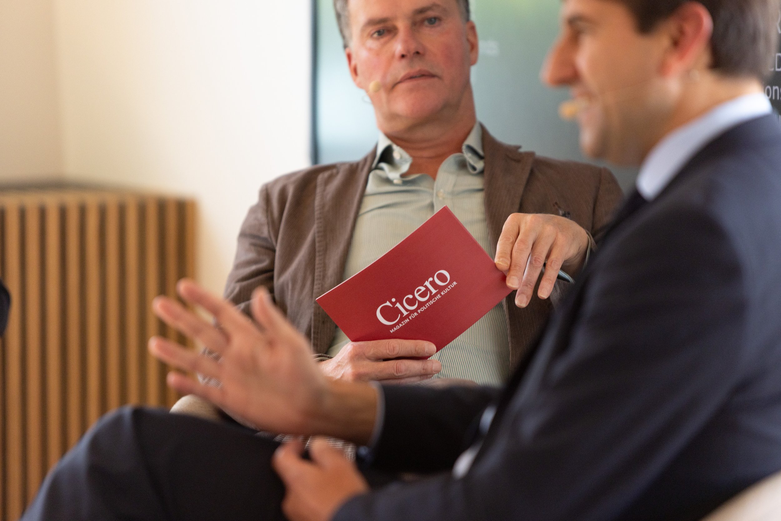 Zwei Männer im Gespräch, einer hält ein rotes Magazin mit dem Titel "Cicero" sichtbar. Der Mann im Hintergrund trägt ein beige Jacket und ein Hemd, der Mann im Vordergrund trägt einen dunklen Anzug.