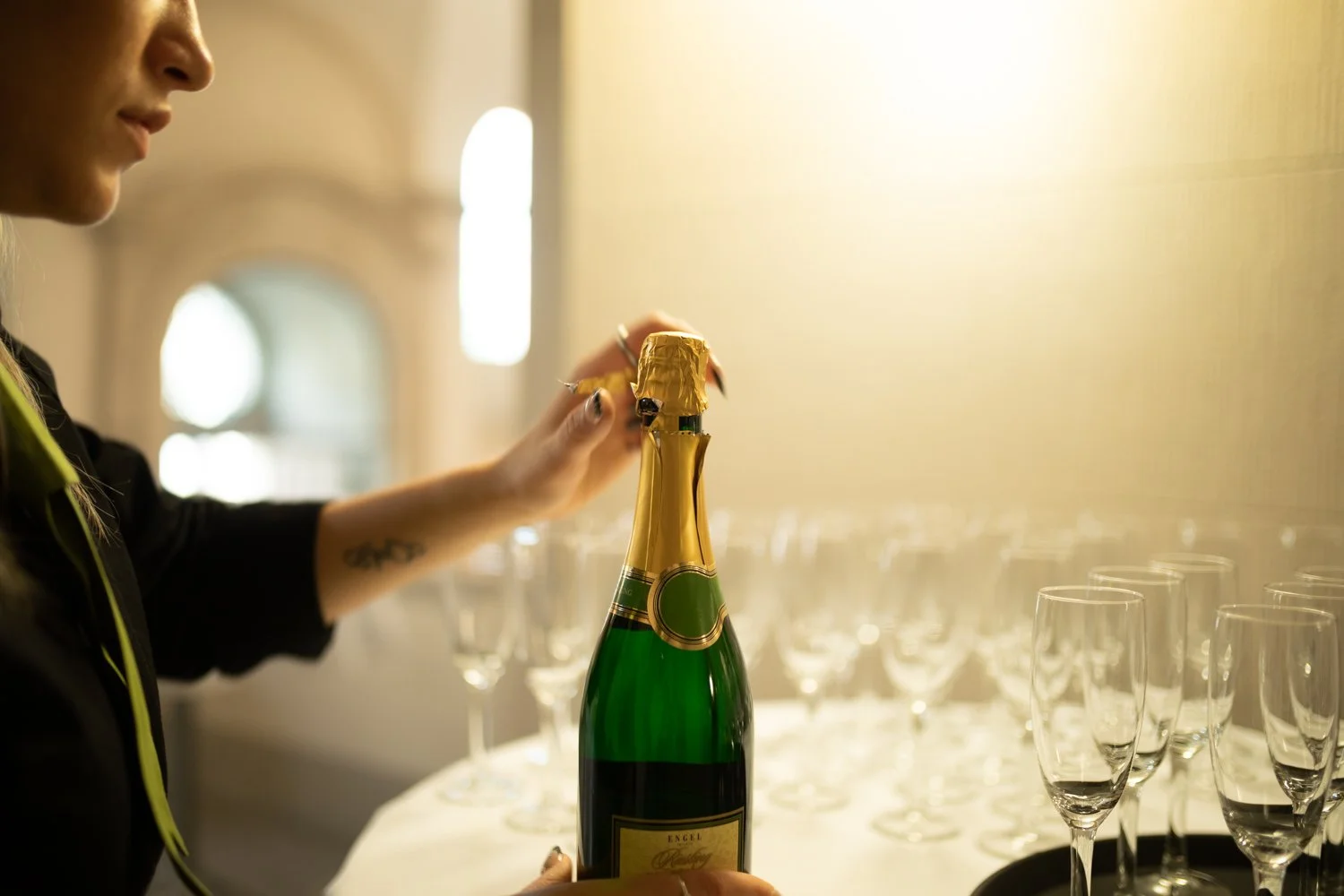 Eine Person öffnet eine Flasche Sekt oder Champagner in einem Raum mit vielen leeren Gläsern auf einem Tisch.