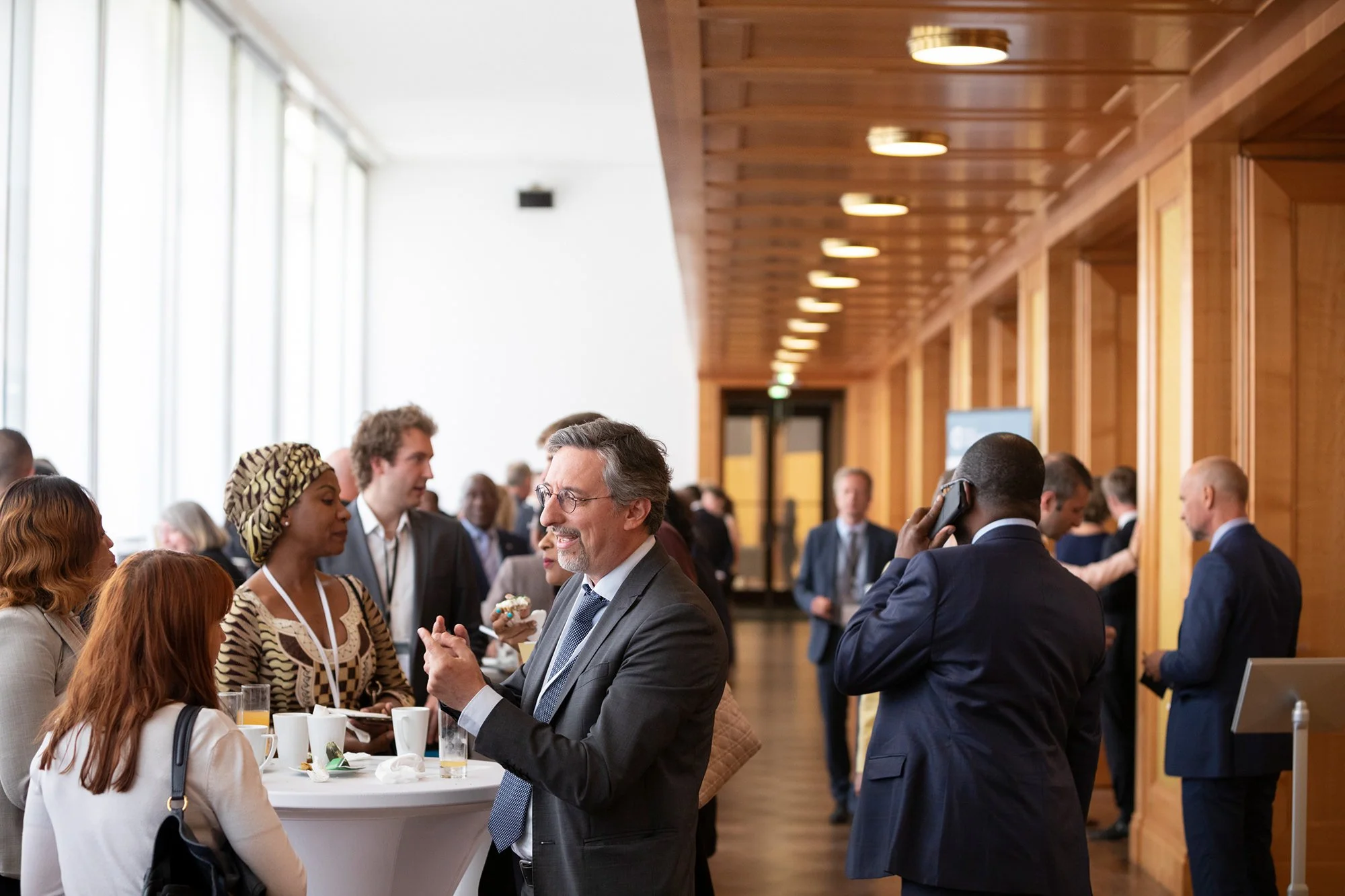 Geschäftsleute bei einem Networking-Event, einige im Gespräch, andere warten oder telefonieren, in einem Raum mit Holzdecke und großen Fenstern.