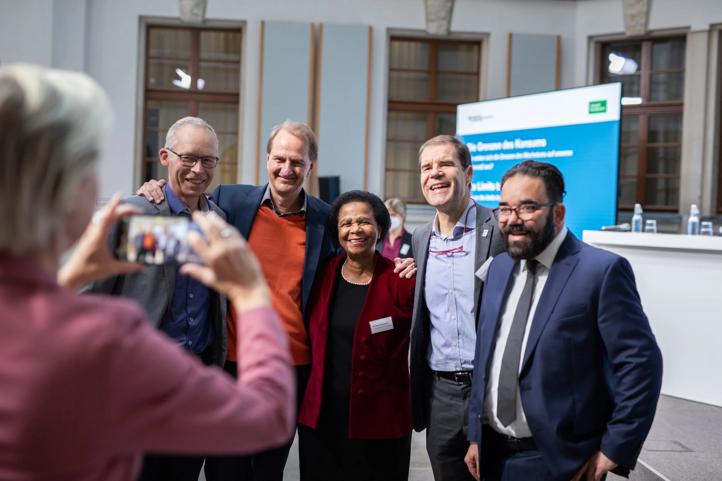 Gruppe von lachenden Menschen, die für ein Foto posieren, während eine Person im Vordergrund ein Smartphone benutzt, um sie zu fotografieren, im Hintergrund ein Bildschirm mit Text und Wasserflaschen auf einem Tisch.
