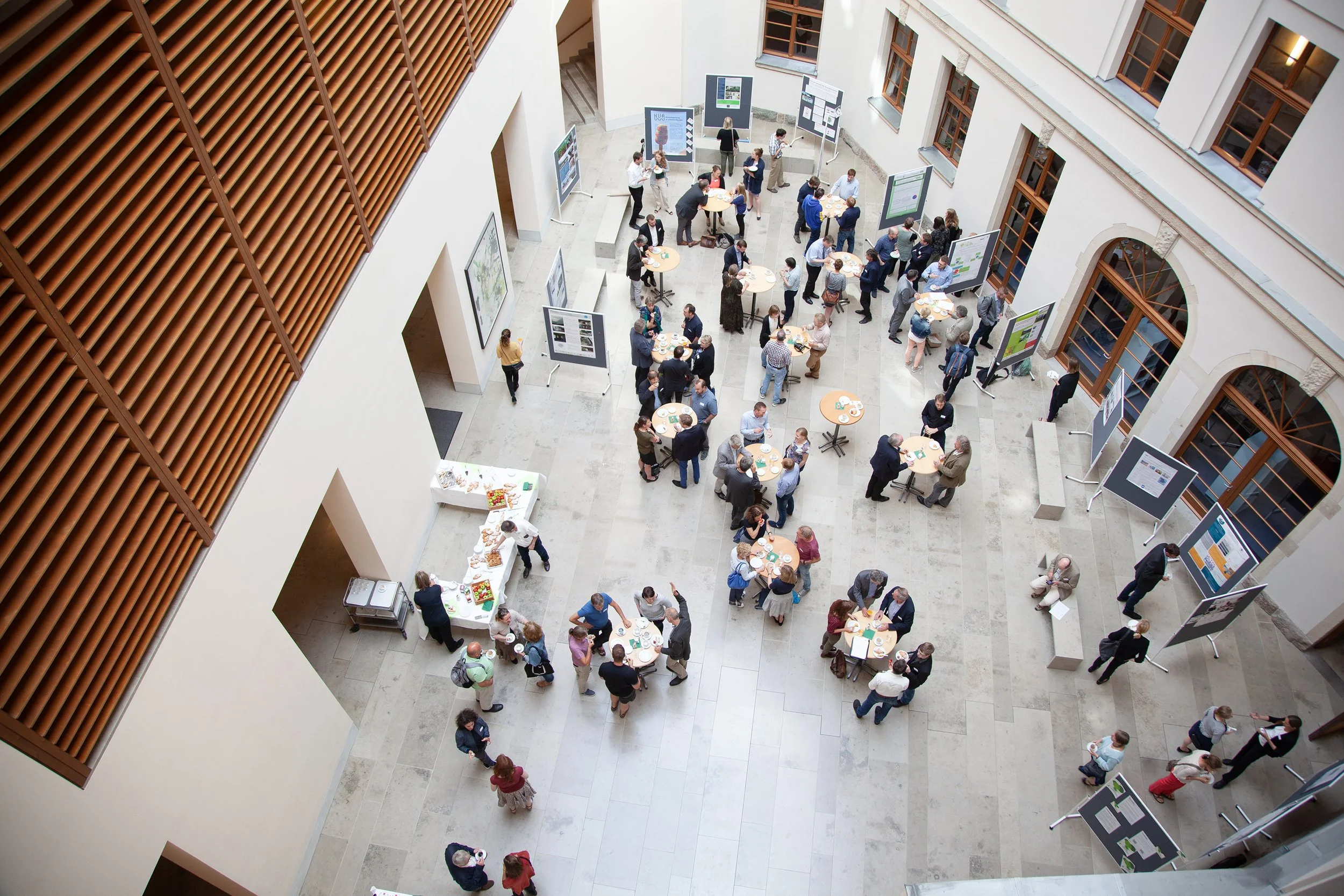 Blick in einen hellen Innenhof eines historischen Gebäudes mit großen Fenstern und Holzverkleidung. In der Mitte sind Menschen bei einer Ausstellung oder Konferenz mit Tischen, Postern und mehreren kleinen Gruppen im Gespräch.