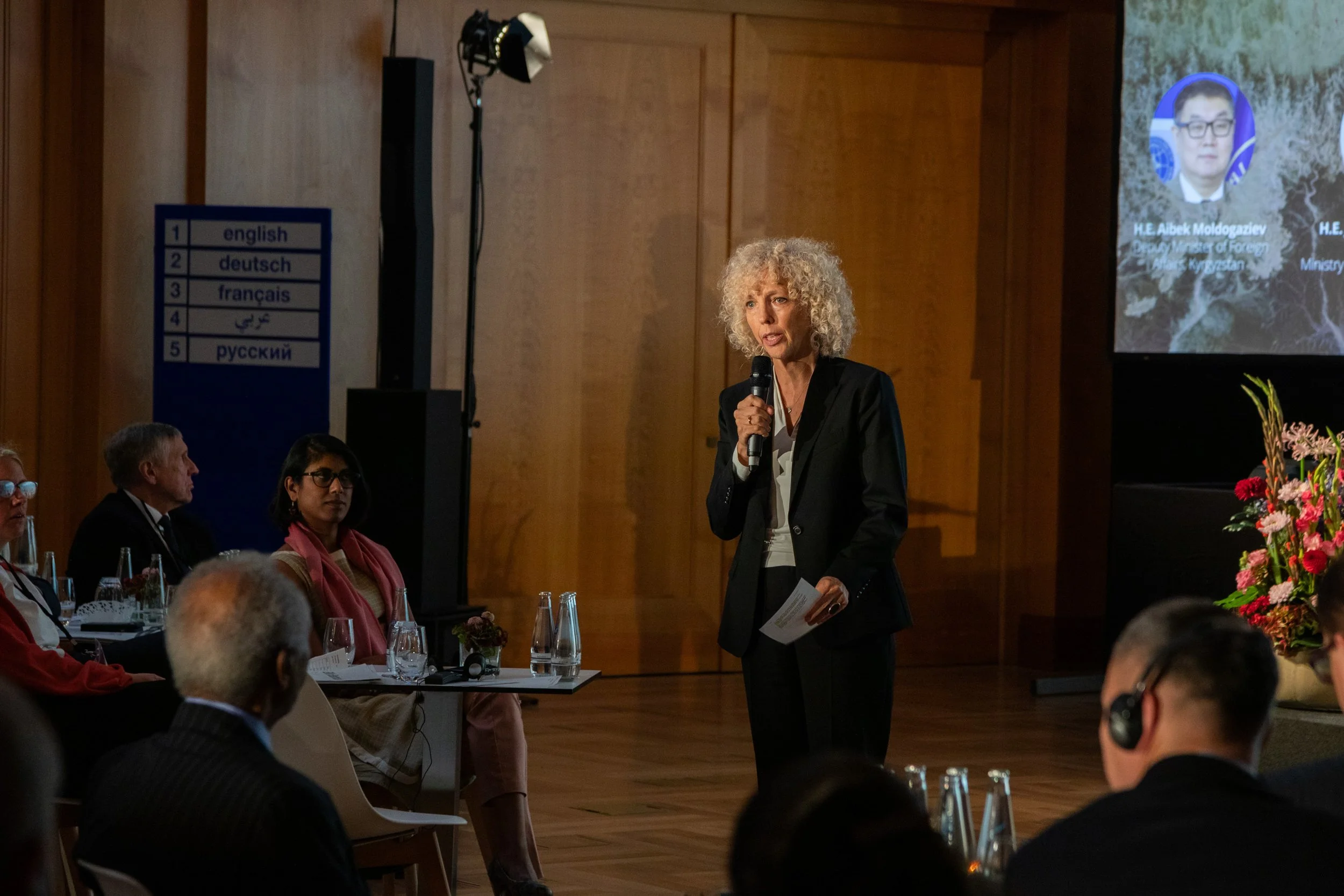 Eine blonde Frau hält ein Mikrofon und spricht bei einer Veranstaltung vor Publikum. Im Hintergrund ist eine große Projektionsleinwand mit einem Porträt von H.E. Albek Moldogaziev, Minister für Umwelt, und ein Tisch mit mehreren Wasserflaschen und Bl