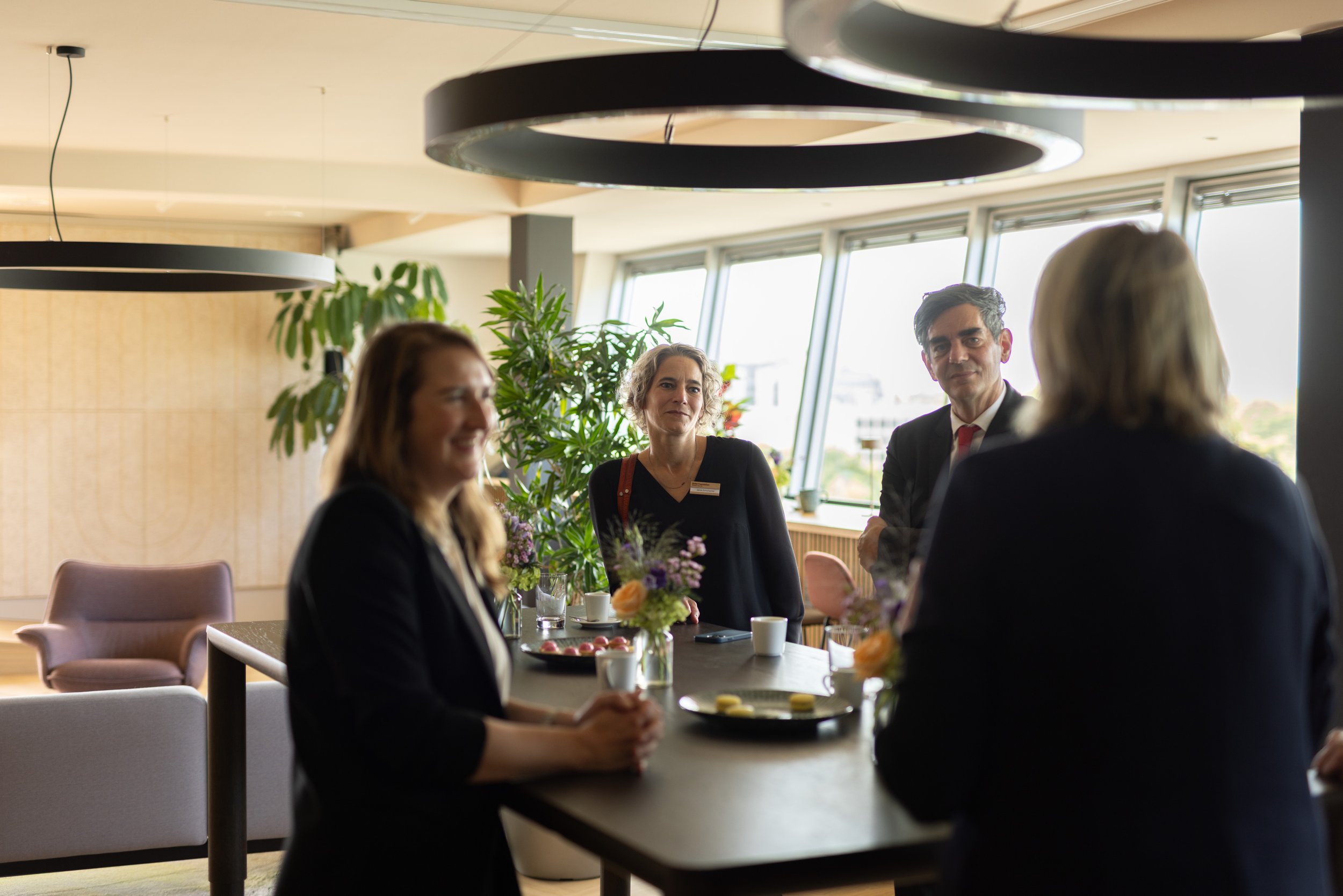 Vier Menschen sitzen in einem modern eingerichteten Raum an einem Tisch bei einer Diskussion oder einem Treffen, mit Pflanzen im Hintergrund und großen Fenstern, die Tageslicht hereinlassen.