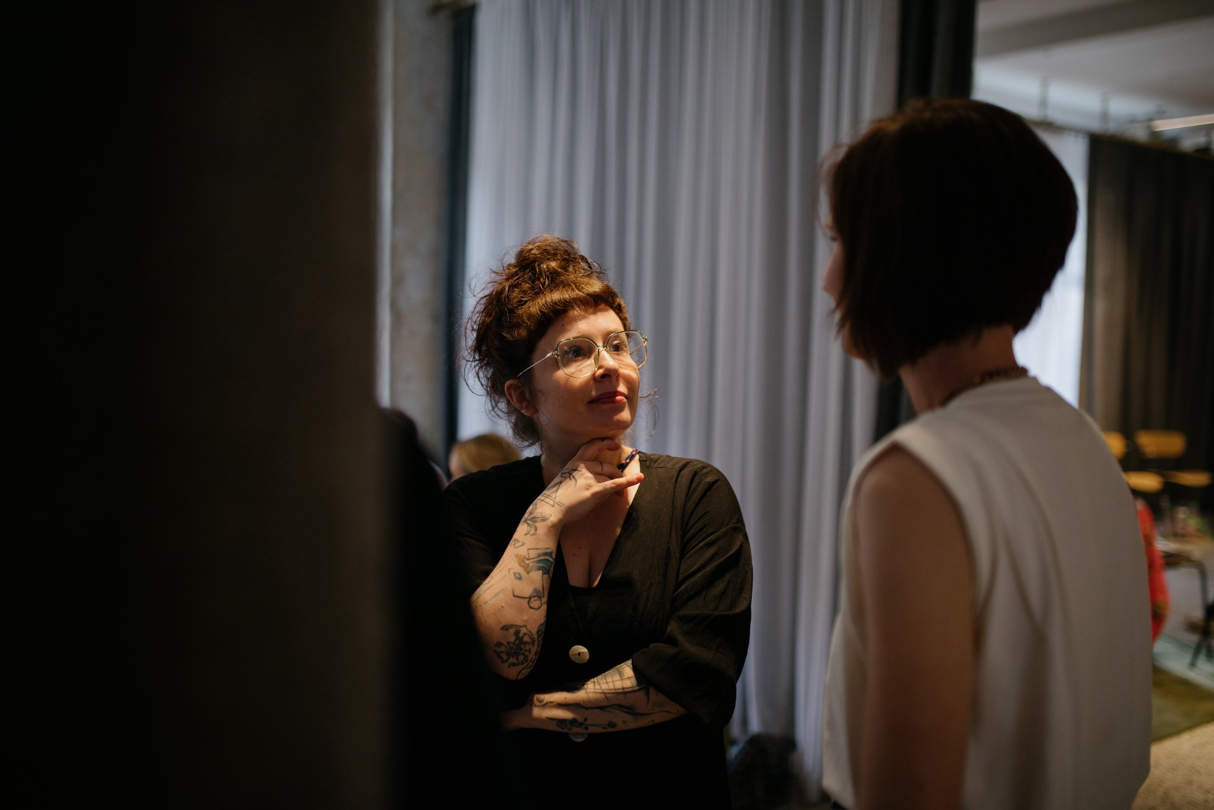 Eine Frau mit roten, lockigen Haaren, Brille und Tattoos an den Armen unterhalten sich mit einer anderen Person.