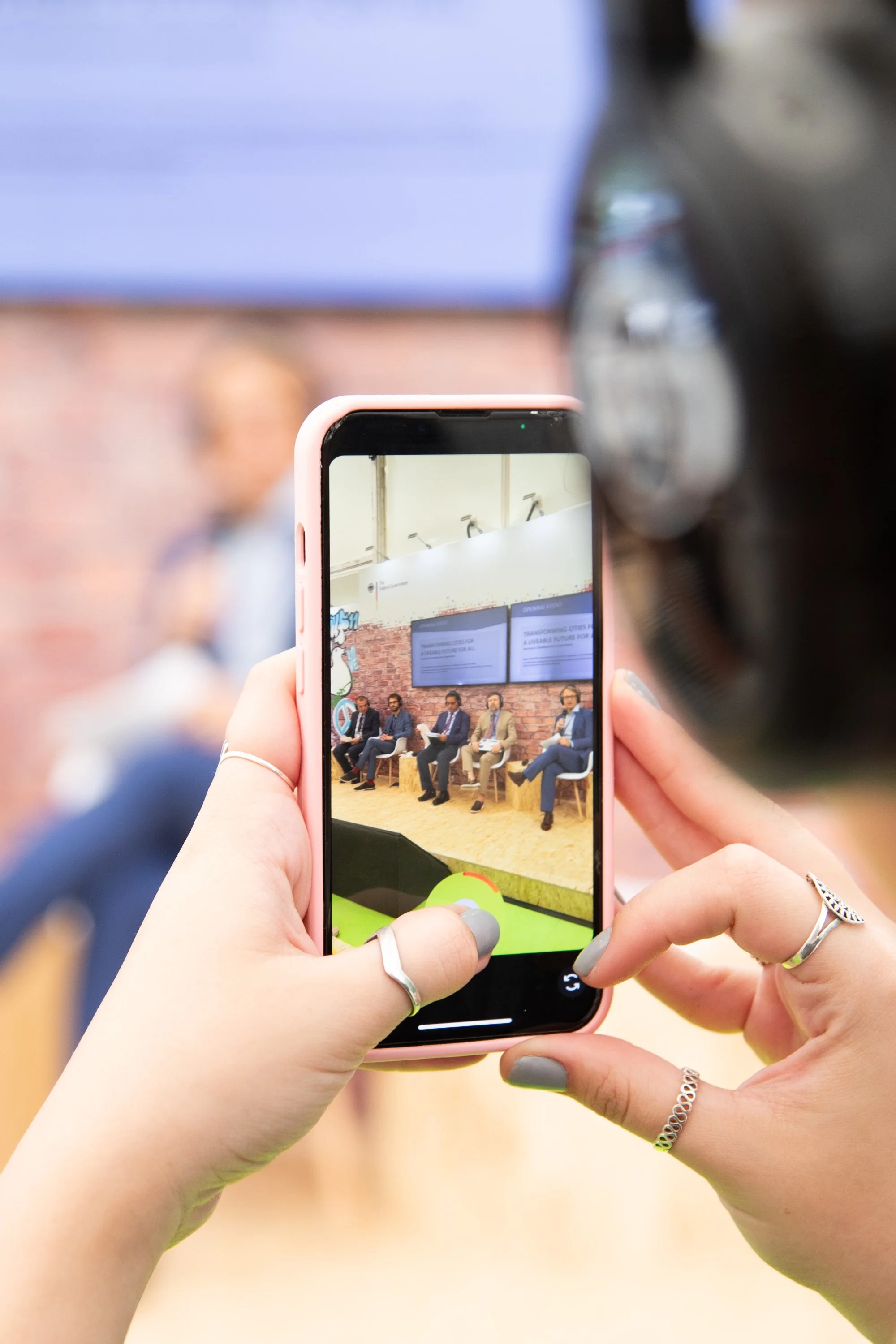 Person hält Smartphone, das ein Panel mit mehreren Sprechern bei einer Veranstaltung zeigt.