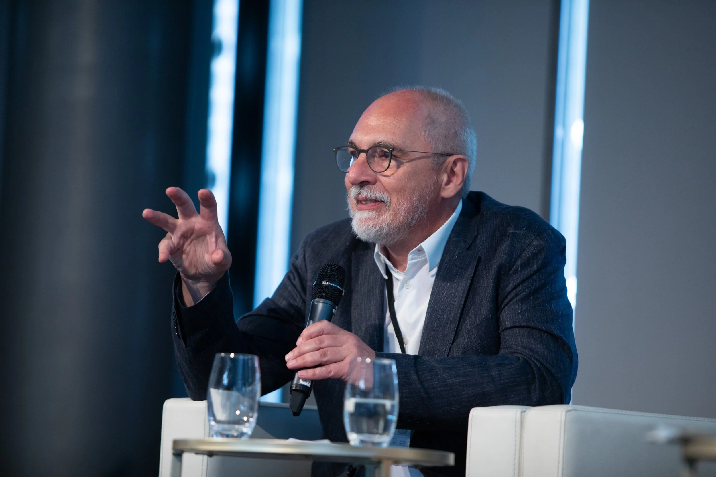 Ein älterer Mann mit Brille hält ein Mikrofon und spricht auf einer Konferenz oder einem Panel. Er trägt einen dunklen Anzug und sitzt an einem weißen Tisch, auf dem zwei Gläser Wasser stehen, während im Hintergrund vertikale blaue Lichter leuchten.