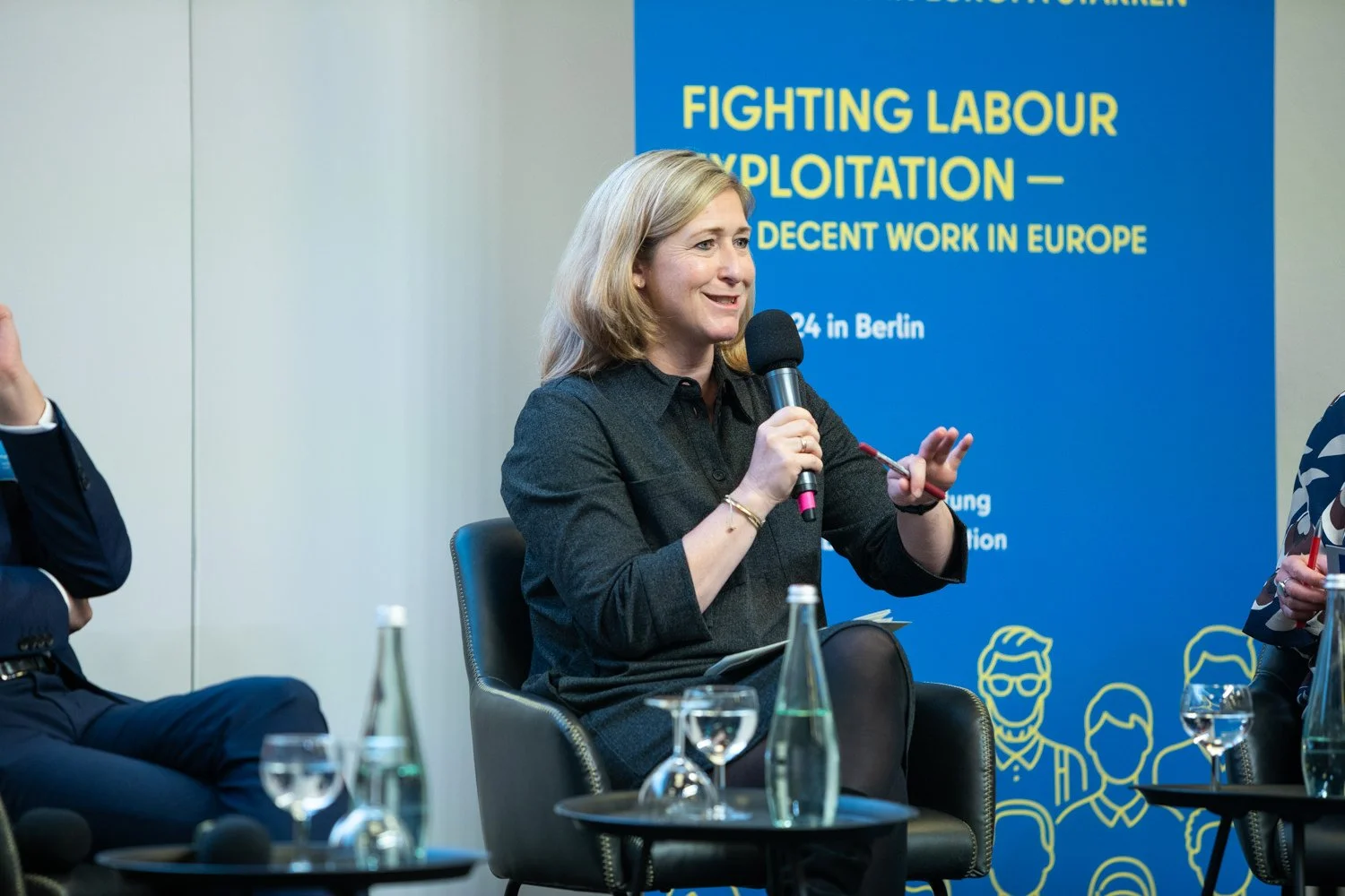 Eine Frau mit blonden Haaren hält einen Mikrofon und spricht bei einer Podiumsdiskussion. Im Hintergrund ist ein blauer Banner mit gelbem Text zu sehen, der das Thema 'Kämpfende Arbeitsausbeutung – Würdige Arbeit in Europa' behandelt, mit weiteren In