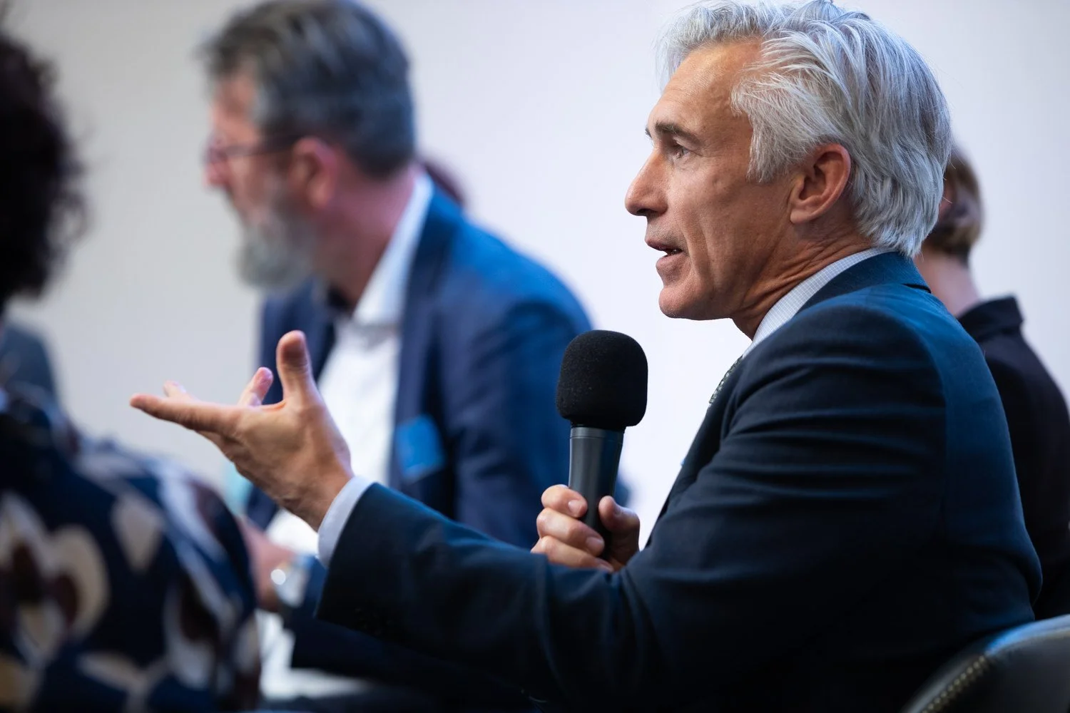 Ein älterer Mann mit weißen Haaren, der einen schwarzen Mikrofon hält und spricht, während er an einem Tisch sitzt, im Gespräch mit anderen Personen, im Hintergrund ablenkbar.