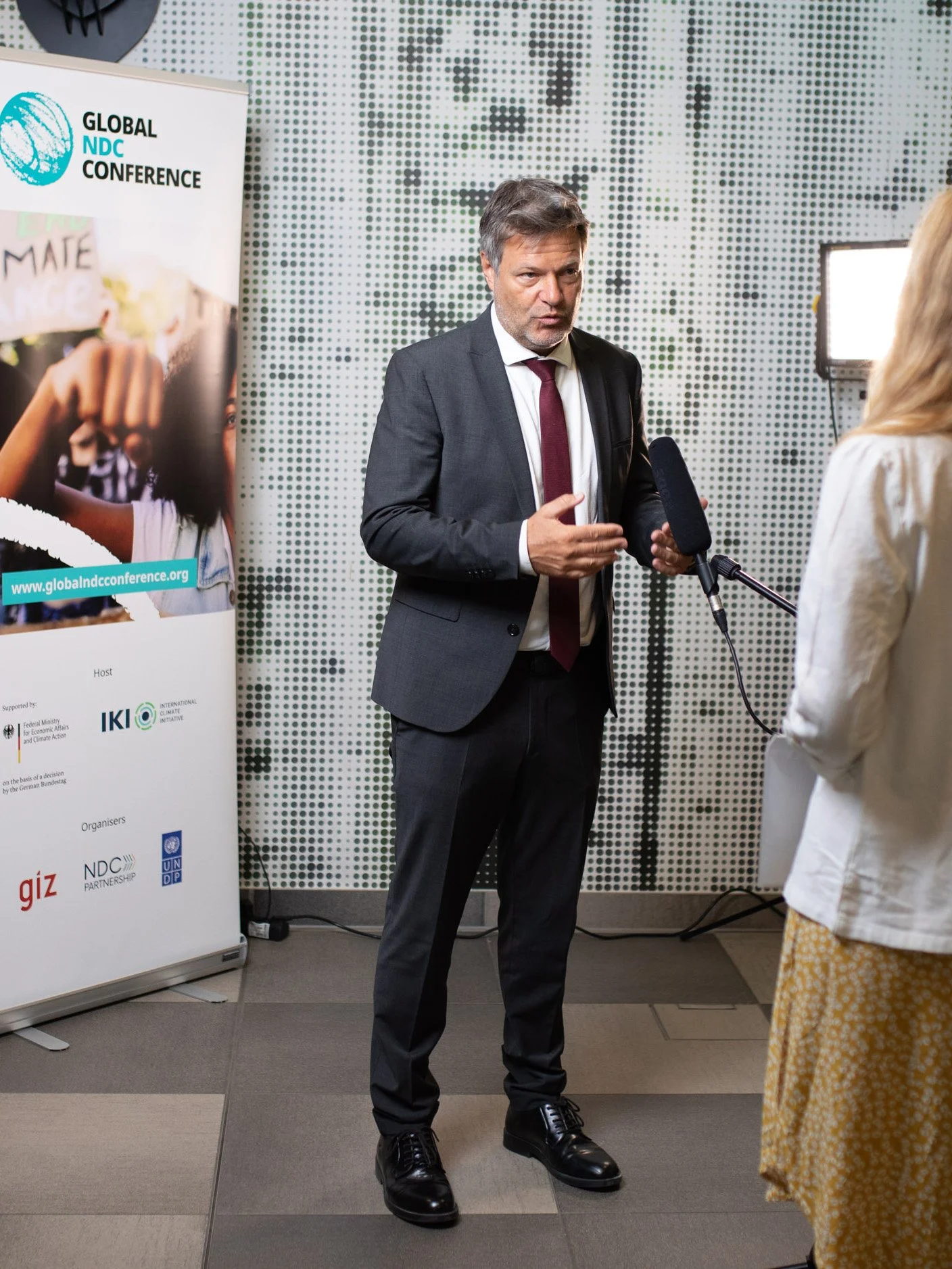 Wirtschaftsminister Robert Habeck wird von einer Frau im Gespräch interviewt. Im Hintergrund ein Plakat mit dem Titel 'Global NDC Conference' und einem Globus-Symbol.