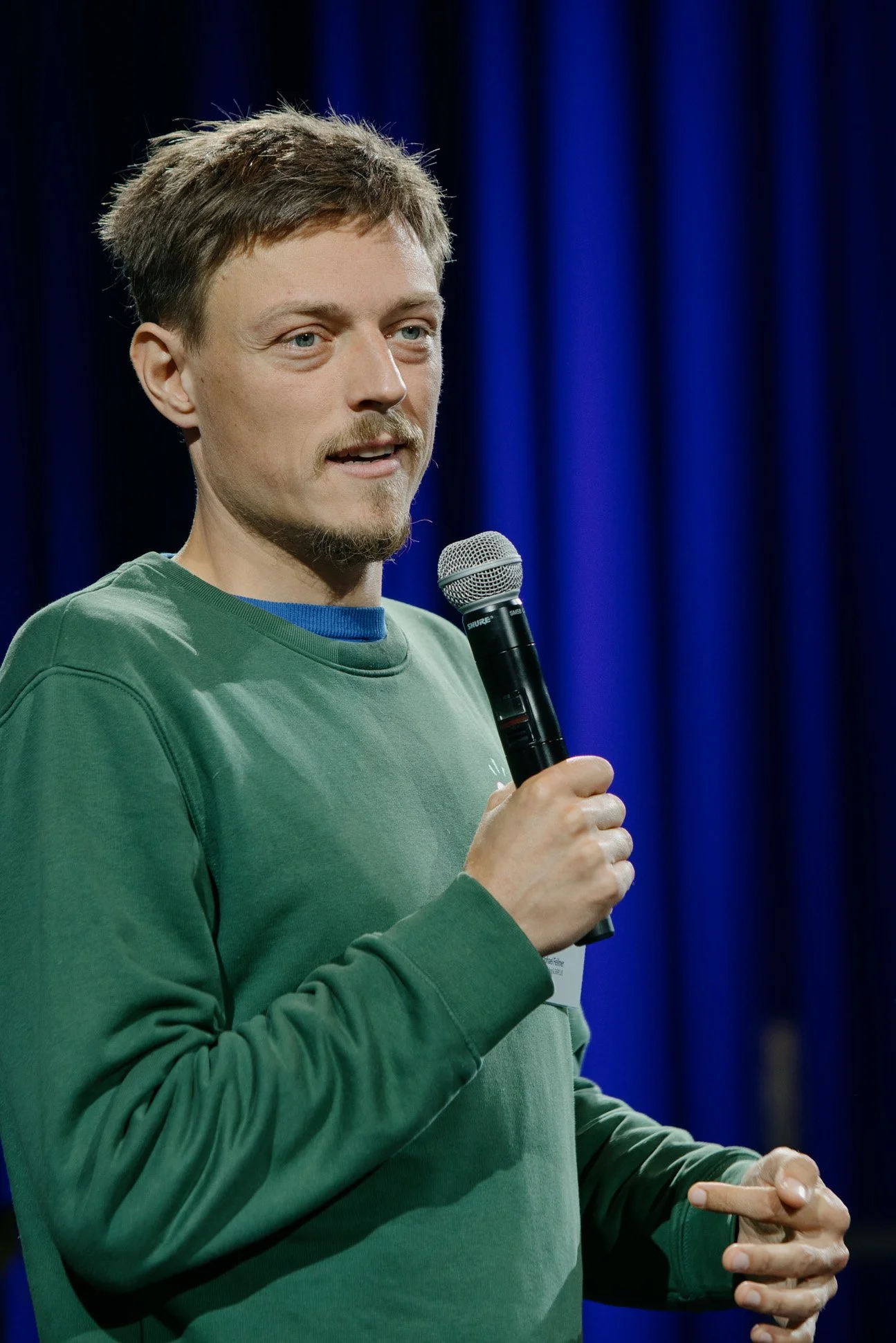 Ein Mann mit braunem Haar, rasiertem Gesicht und blauen Augen hält ein Mikrofon in der Hand und spricht vor einem dunklen blauen Vorhang.