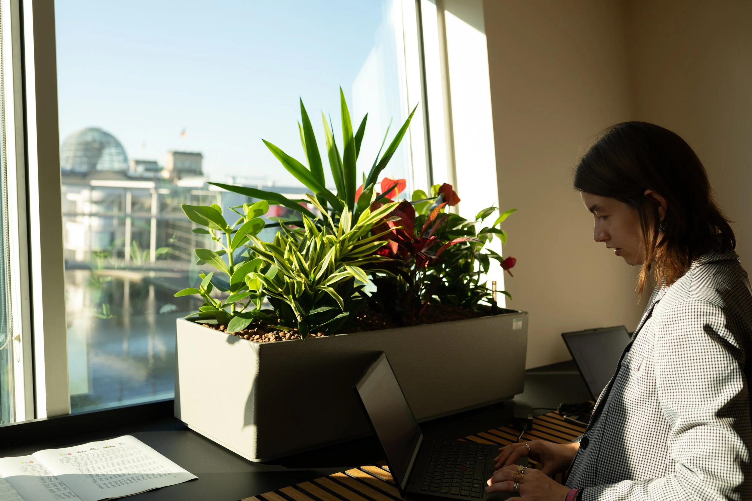 Eine Frau arbeitet an einem Laptop an einem Schreibtisch neben einem Fenster. Auf dem Schreibtisch stehen zwei elektronische Geräte und in der Ecke befindet sich eine große Blumenvase mit grünen und roten Pflanzen.