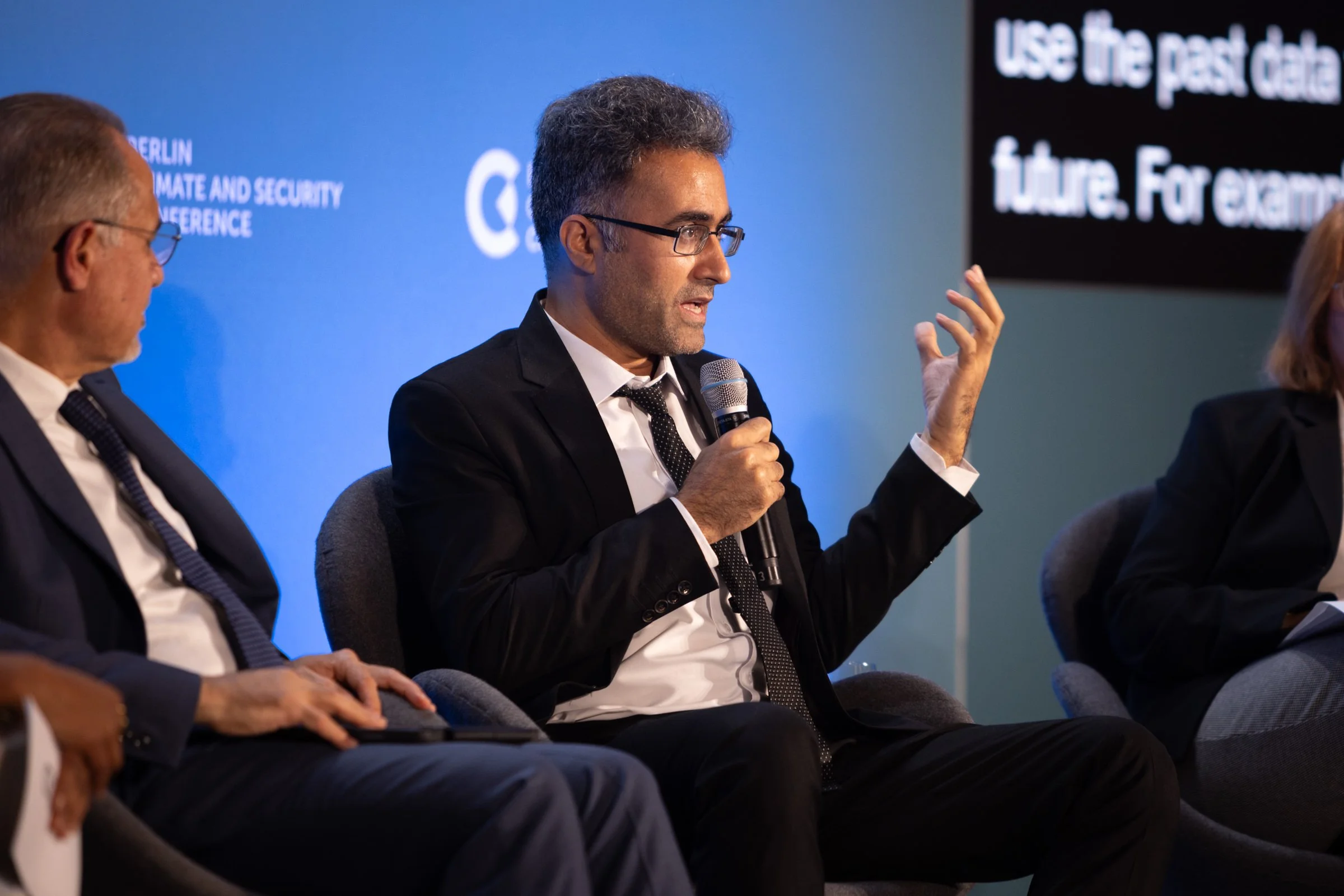 Ein Mann in Anzug und Krawatte spricht bei einer Konferenz und hält ein Mikrofon in der Hand. Im Hintergrund ist ein Bildschirm mit Text zu sehen.