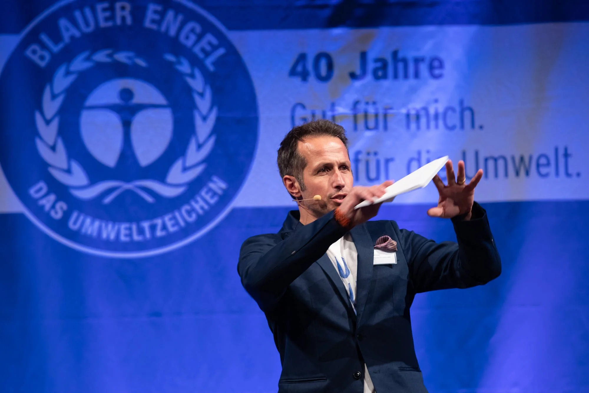 Mann hält ein Blatt Papier in der Hand, spricht bei einer Veranstaltung, im Hintergrund Logo mit Text auf blauer Wand, Feier zum 40-jährigen Bestehen für den Umweltschutz.