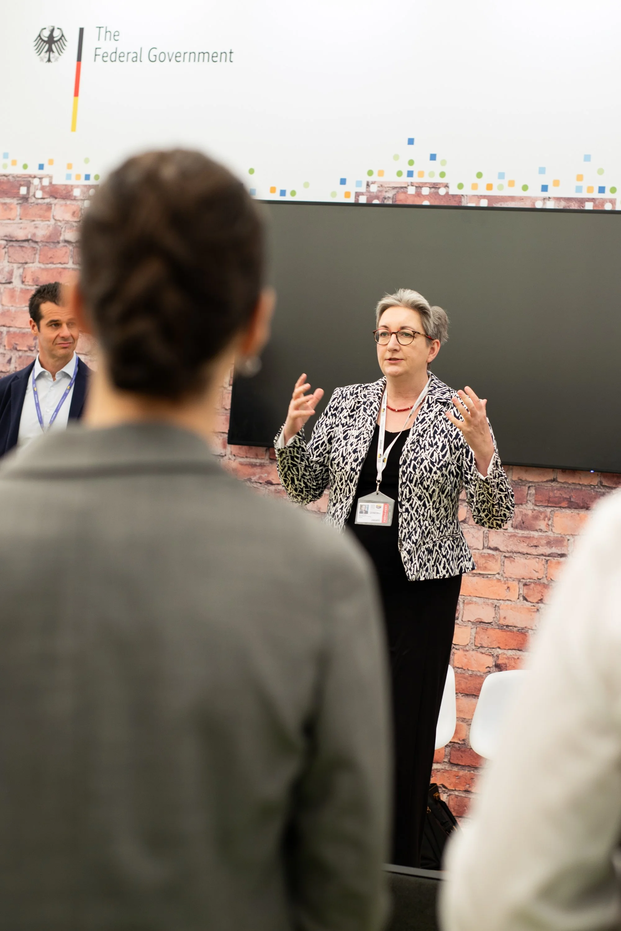 Eine Frau spricht vor einem Publikum bei einer Präsentation, im Hintergrund eine Anzeige des deutschen Bundesministeriums, eine weitere Person steht daneben.